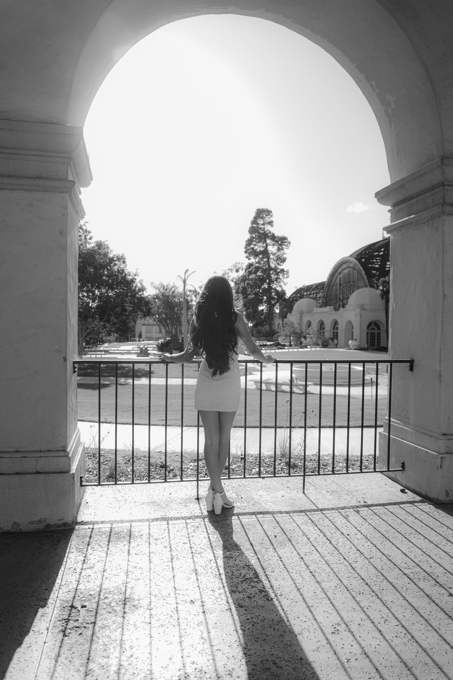 Young woman in white dress stands under an arch overlooking a sunny garden courtyard, black and white photo.