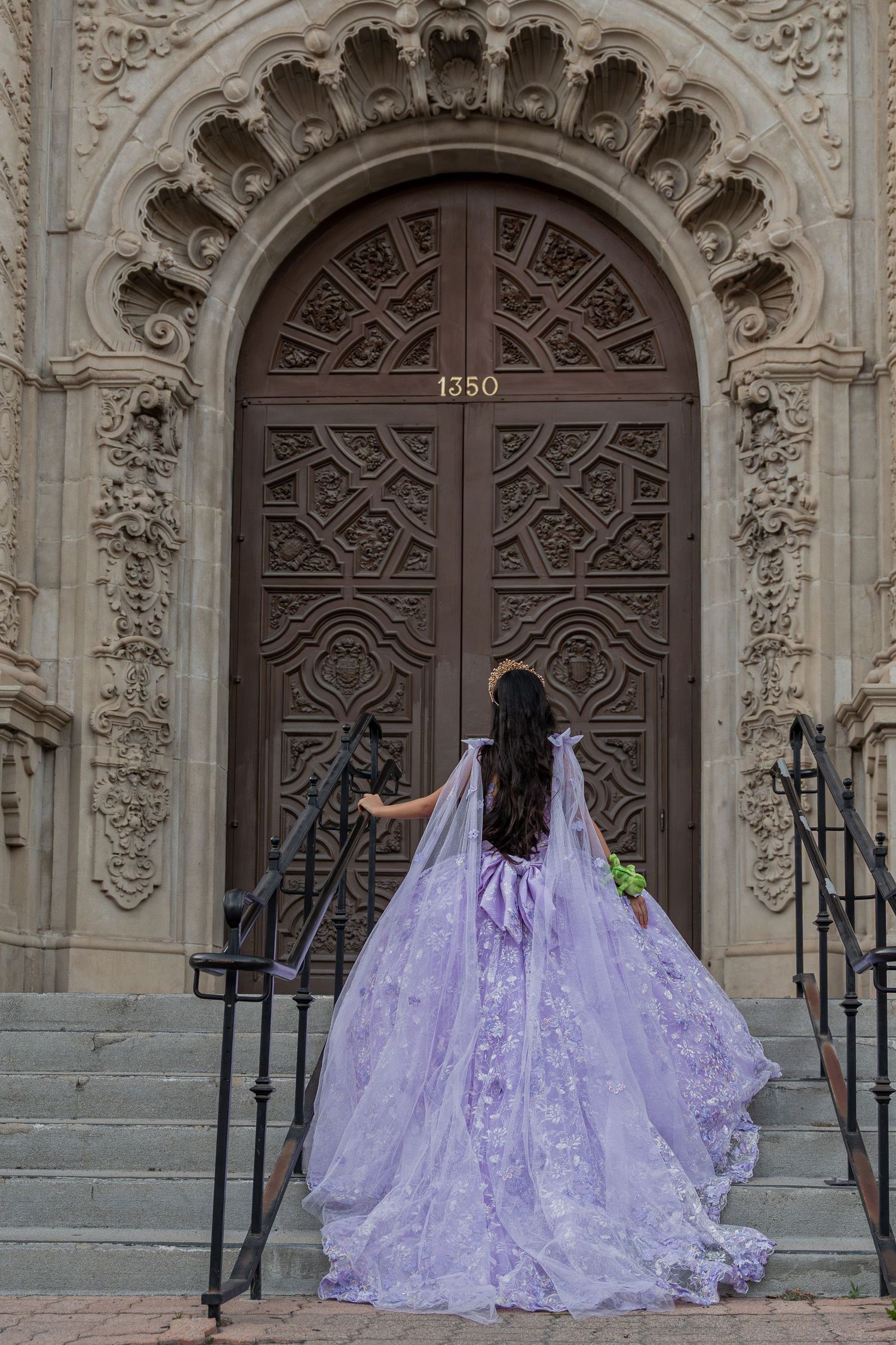 Young woman in lavender quinceañera ball gown with cape stands before ornate carved wooden doors numbered 1350.