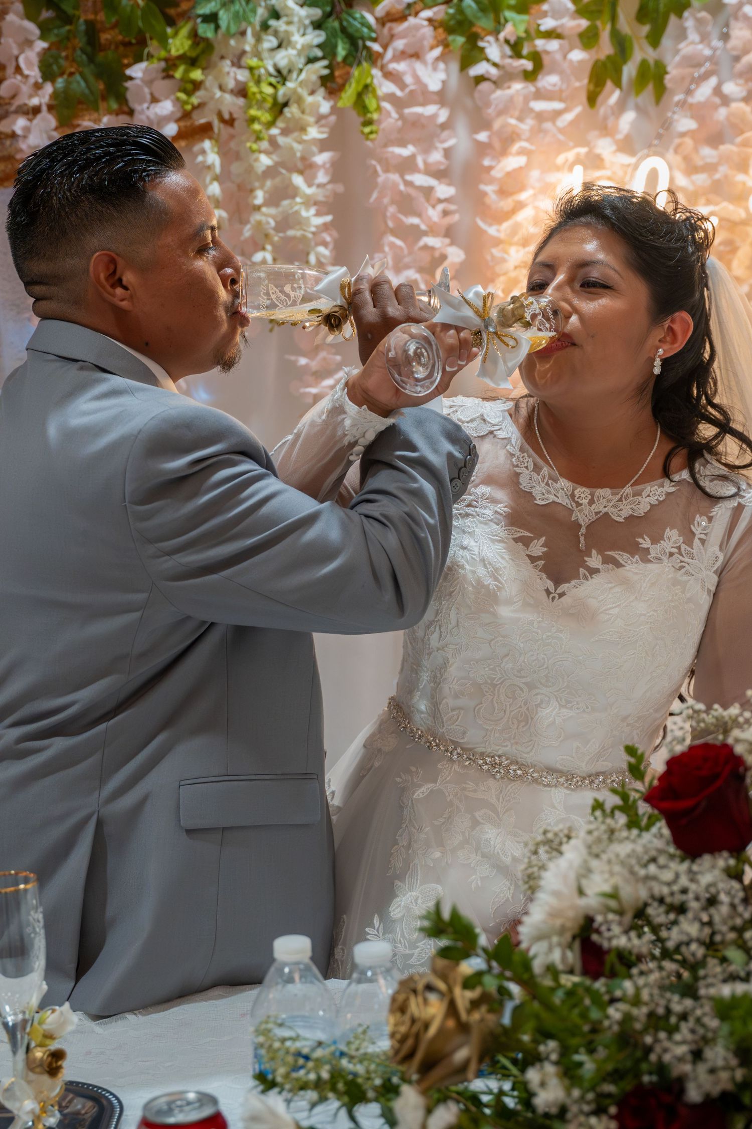 Bride and groom in wedding attire sharing a champagne toast at their reception table with floral decorations.