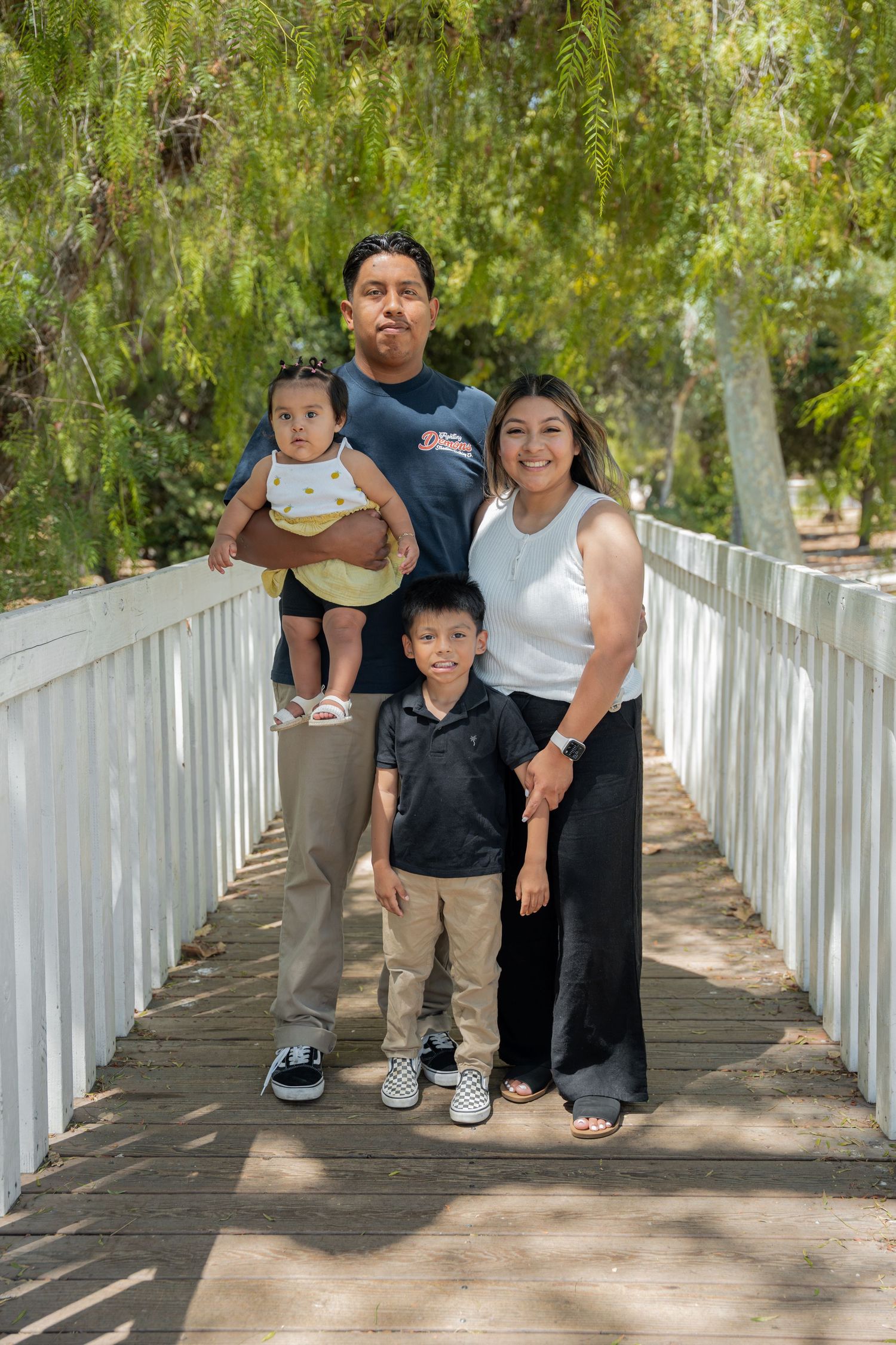 Happy family of four posing together on a white wooden bridge surrounded by green trees in a park.