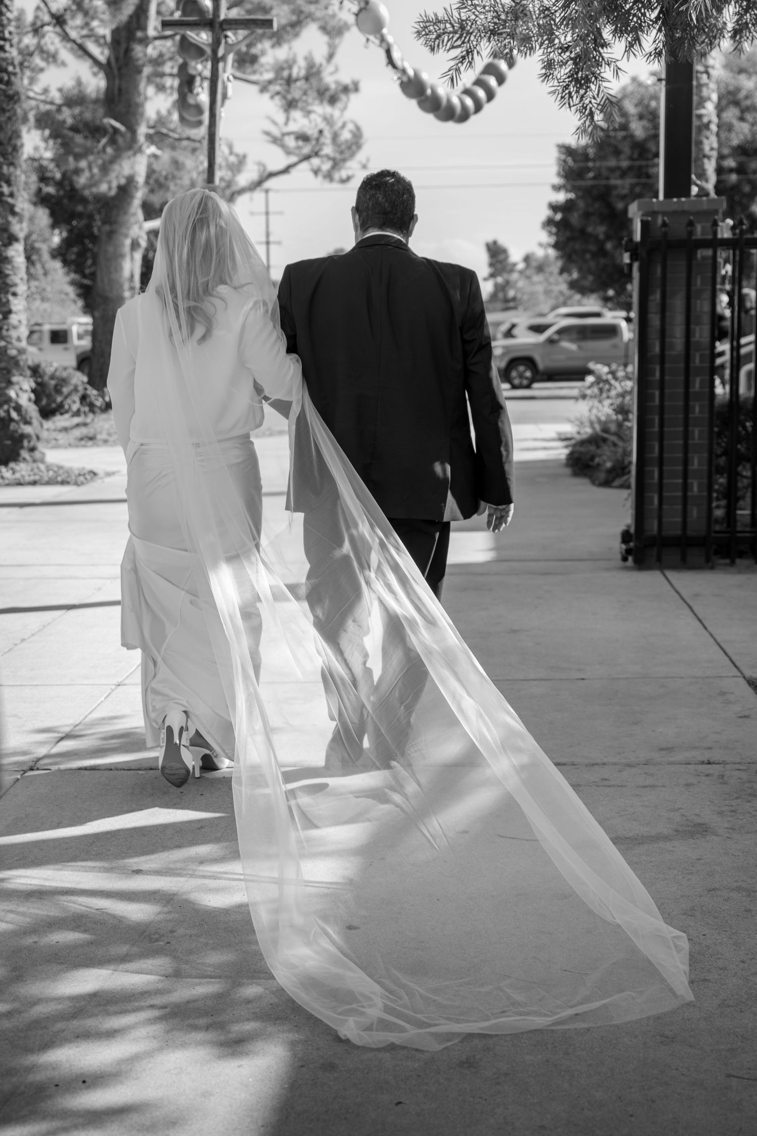 Bride in white gown with long veil walking with groom on sunny sidewalk.