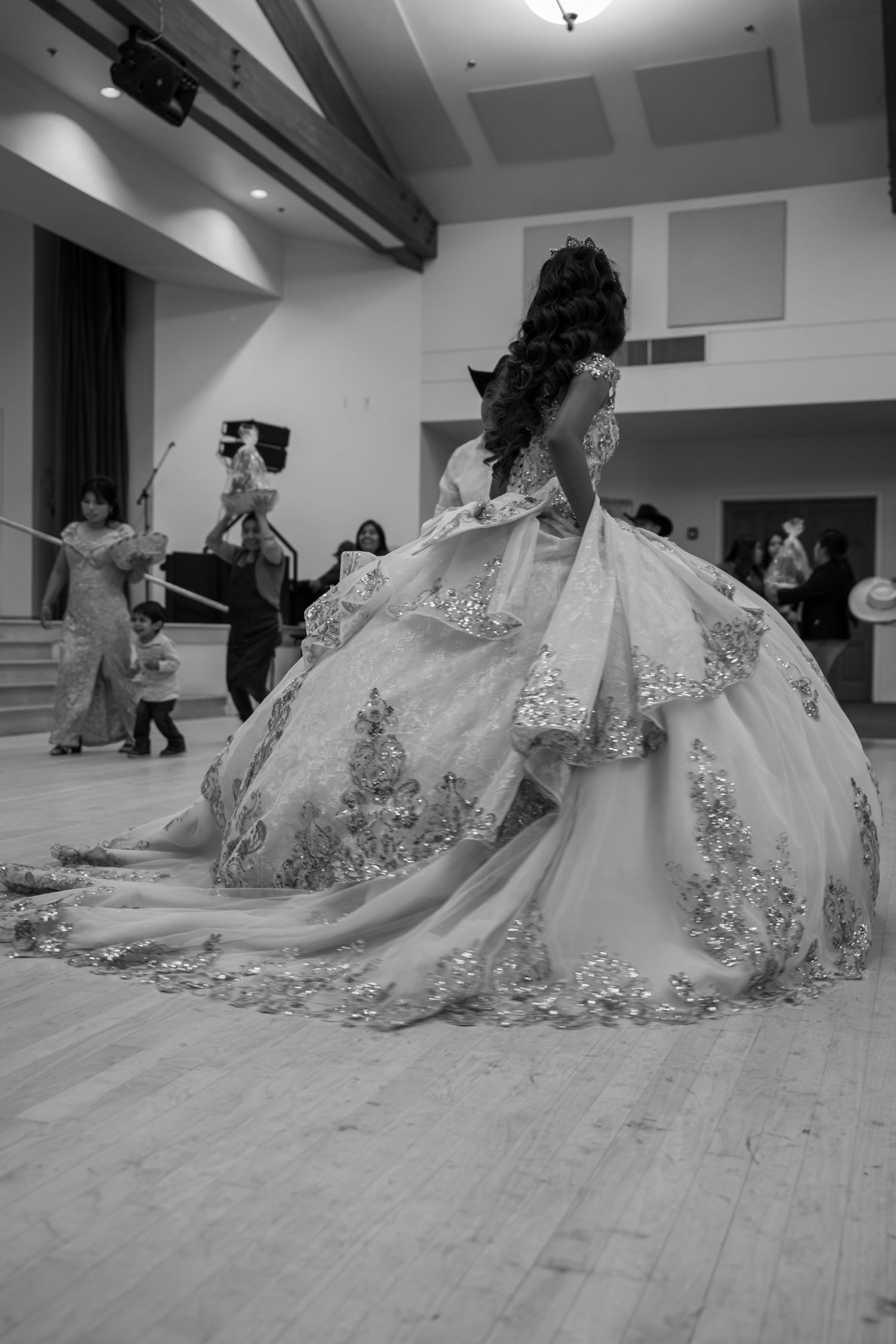 Young woman in stunning white quinceañera gown with gold embroidery poses on dance floor, black and white photo.