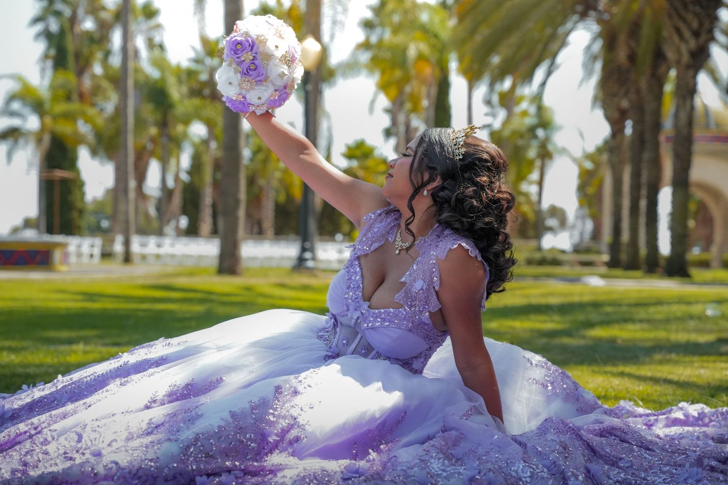 Young woman in a sparkling lavender quinceañera gown seated on grass holding up a bouquet surrounded by palm trees.