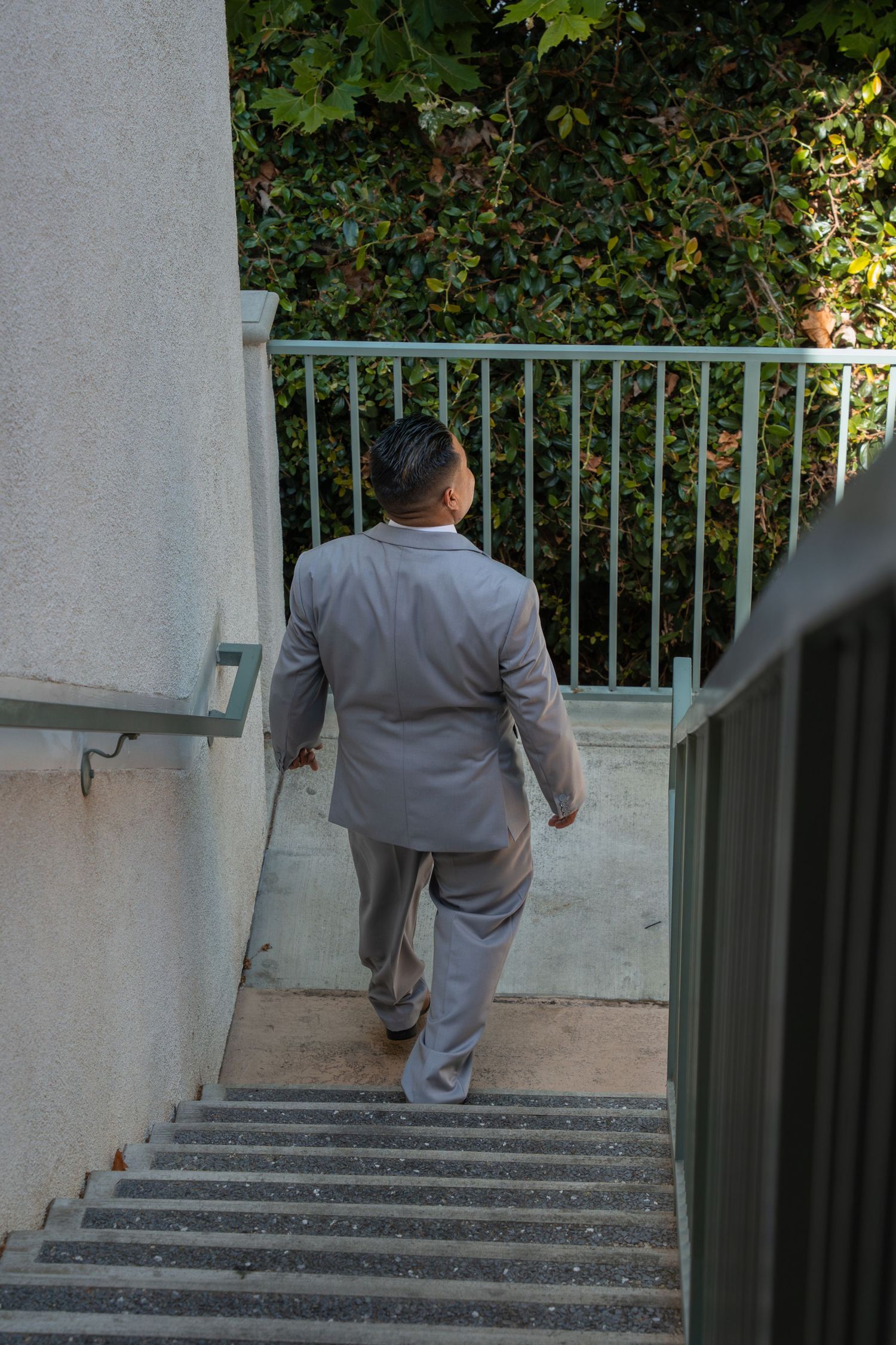Man in a gray suit walking up outdoor stairs next to a metal railing with green foliage in the background.