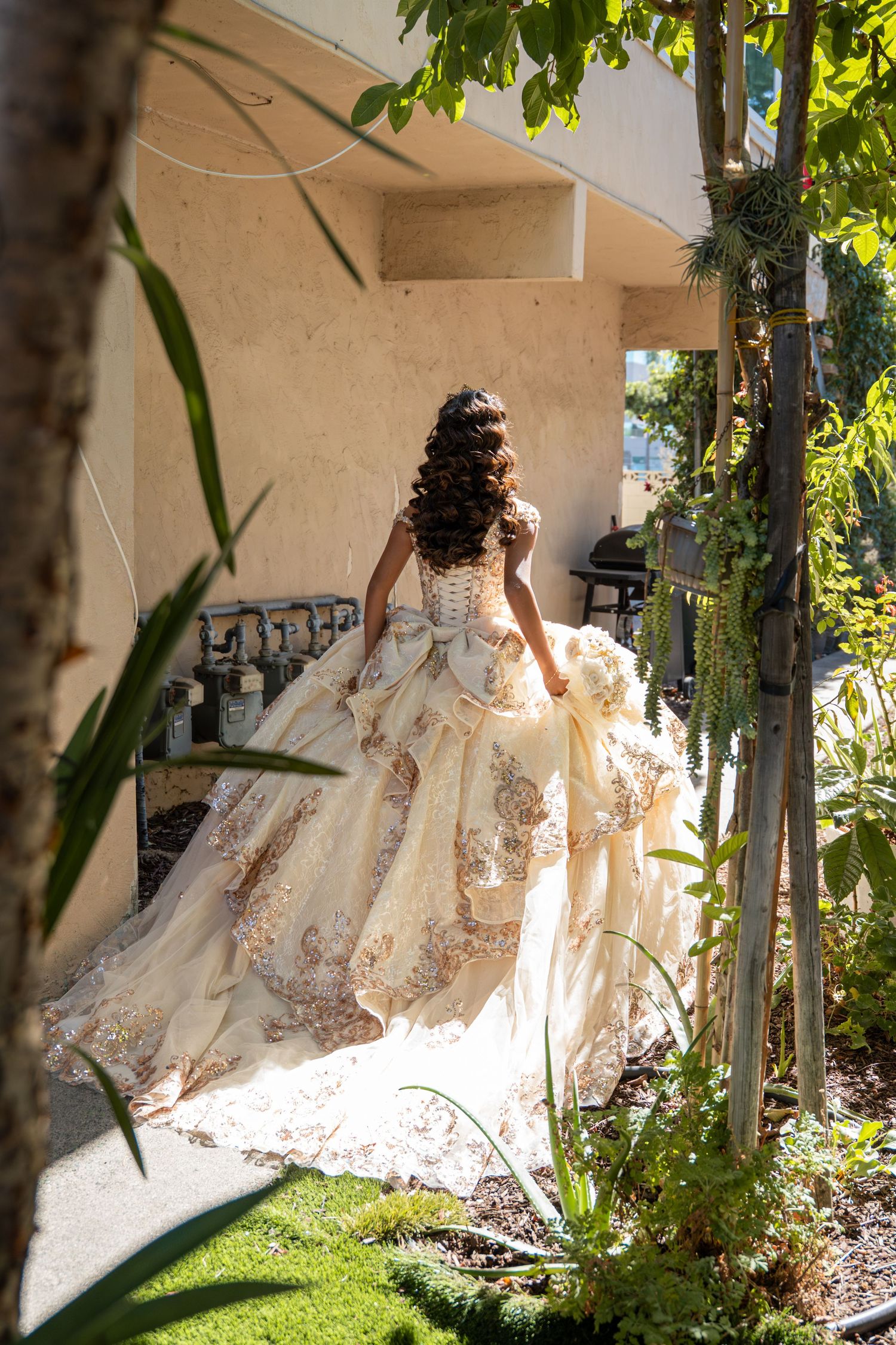 Young woman in ornate gold quinceañera ball gown viewed from behind in a lush garden setting.
