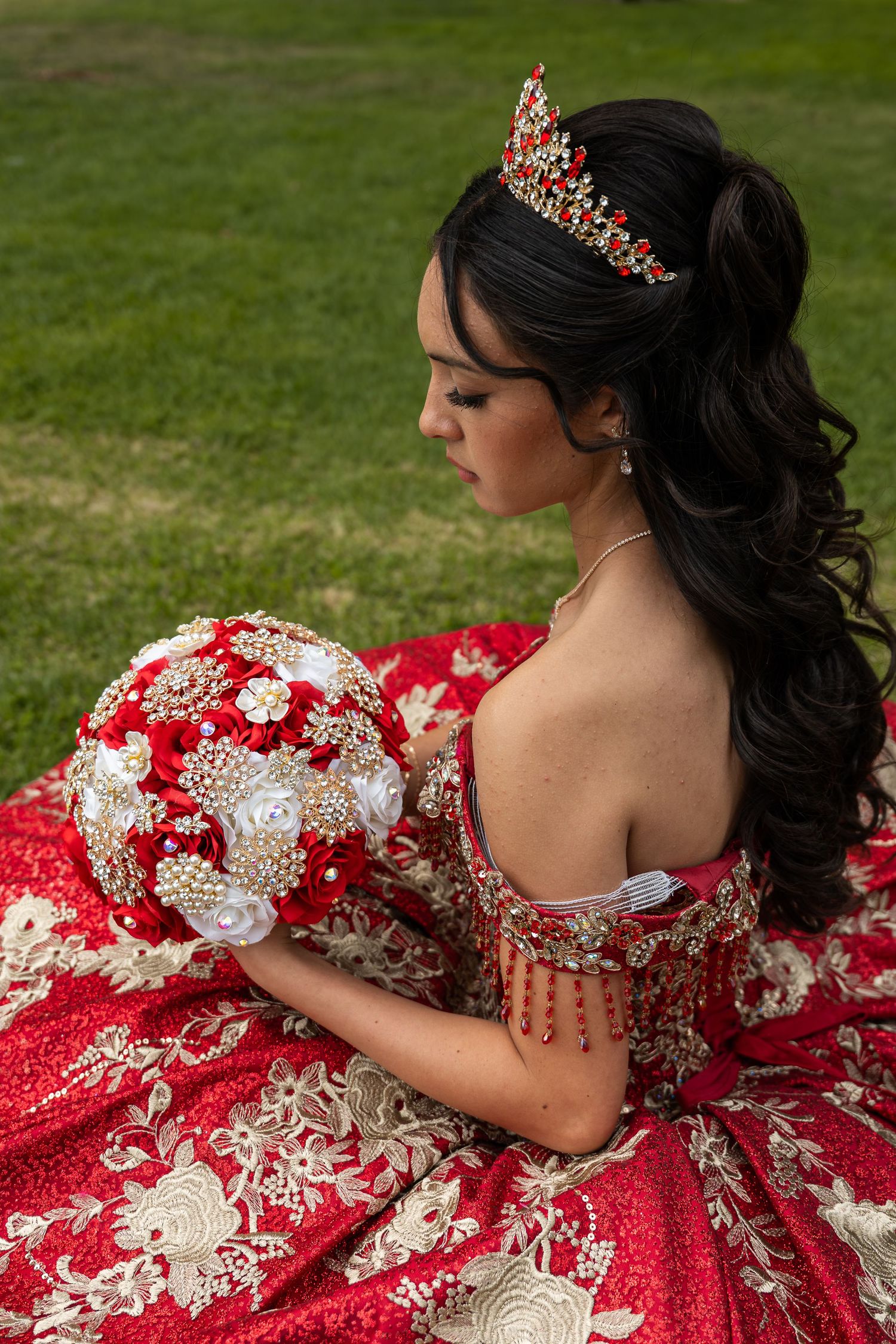 Young woman in red quinceañera gown with gold embroidery, jeweled tiara, and ornate bouquet seated on grass.