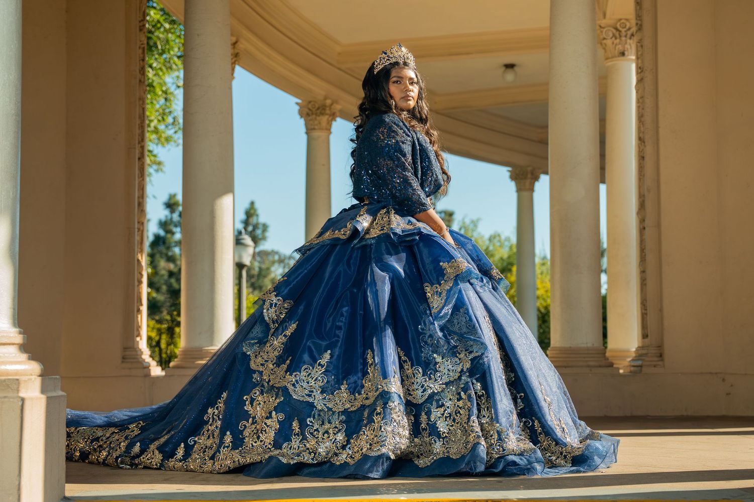Young woman in elegant navy blue quinceañera ball gown with gold embroidery and tiara poses between white columns.