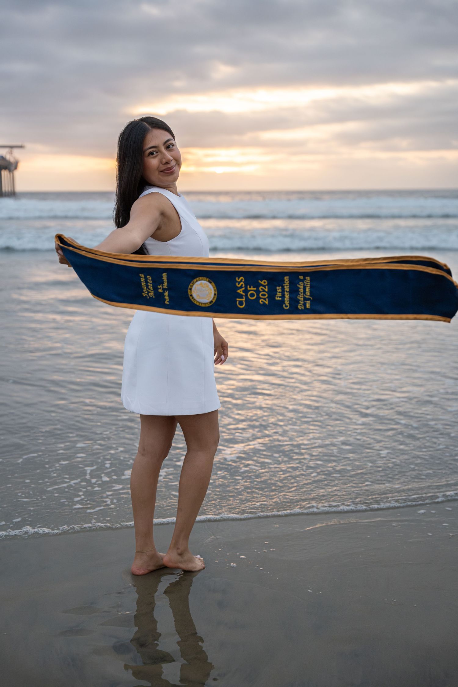 Young woman in white dress holds graduation sash on beach at sunset, celebrating Class of 2025.