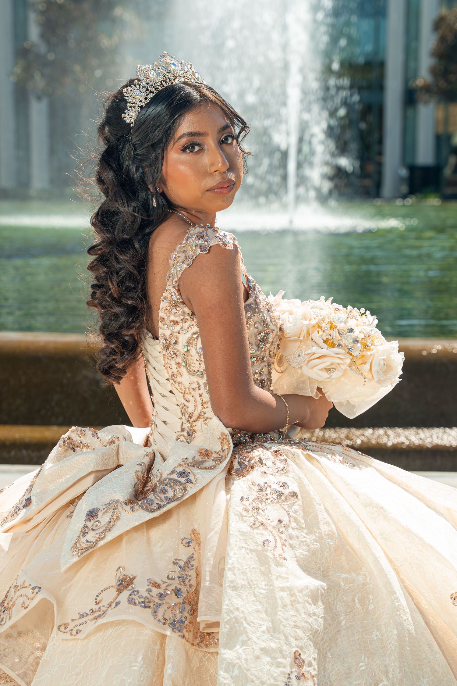 Young Latina woman in elegant champagne quinceañera gown with tiara and bouquet sits by fountain.