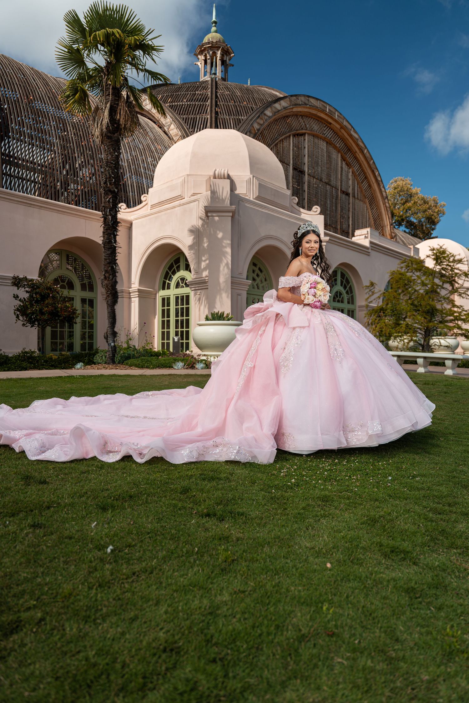 Young woman in a pink ballgown quinceañera dress holding bouquet outside ornate building at Balboa Park.