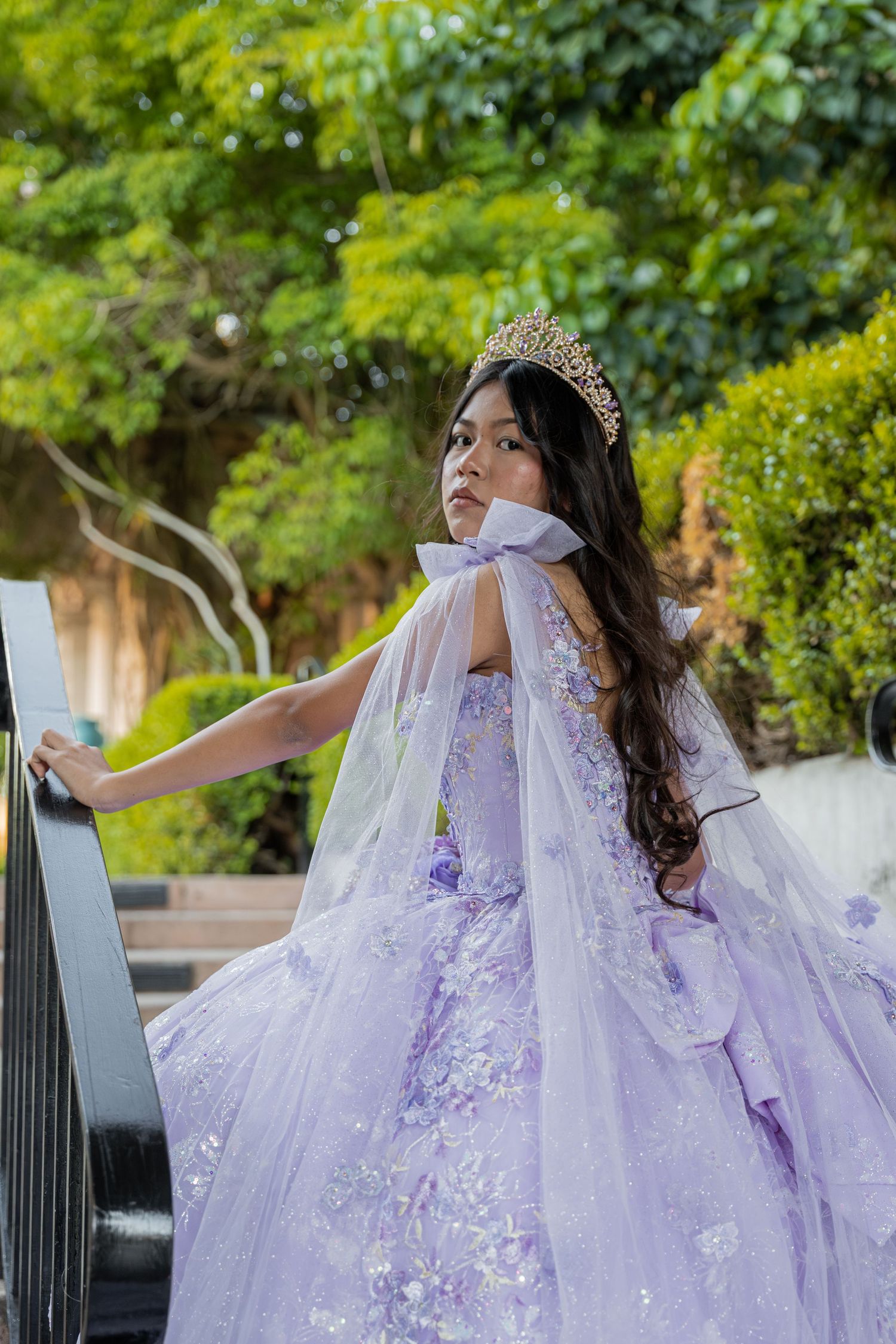 Girl in lavender quinceañera gown with crown and cape posing on outdoor staircase.