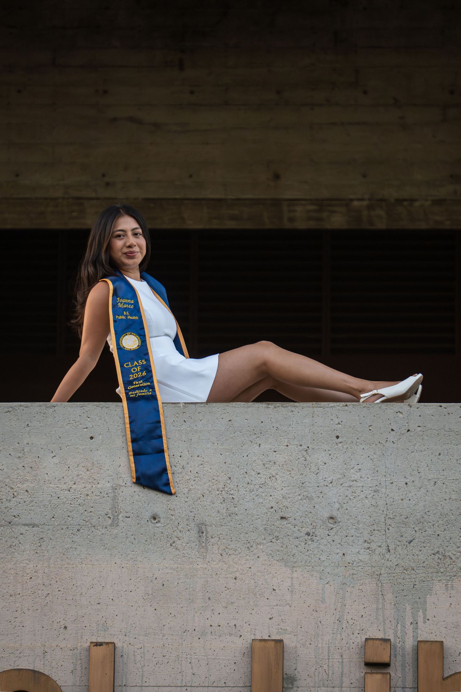 Young woman in white dress and blue graduation sash poses on concrete ledge for graduation photos.