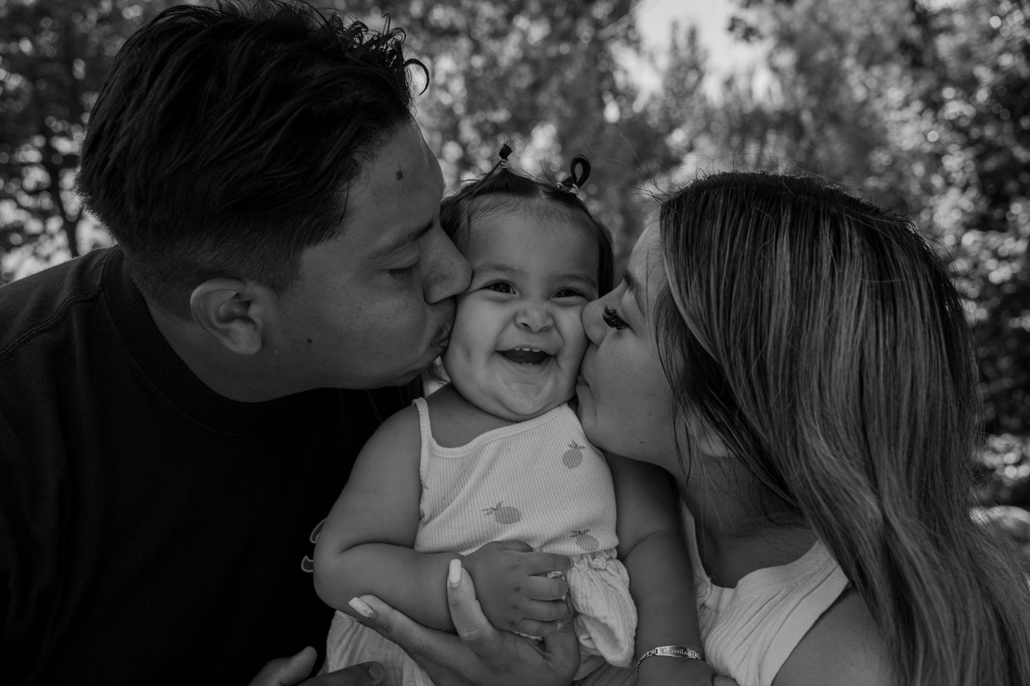 Happy baby laughs joyfully as both parents kiss her cheeks simultaneously in a tender black and white family portrait.
