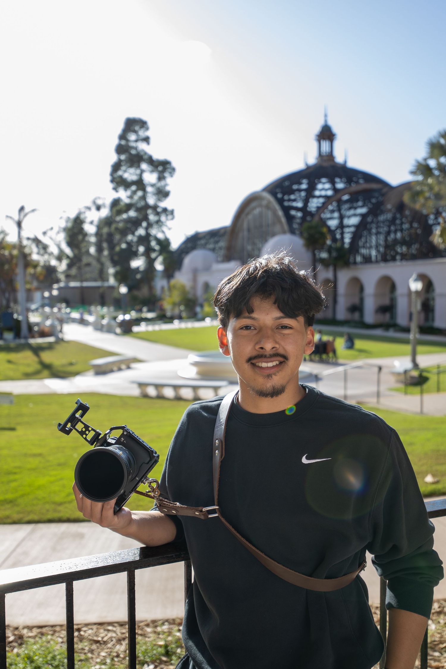 Smiling young male photographer holding professional camera at Balboa Park San Diego on sunny day.