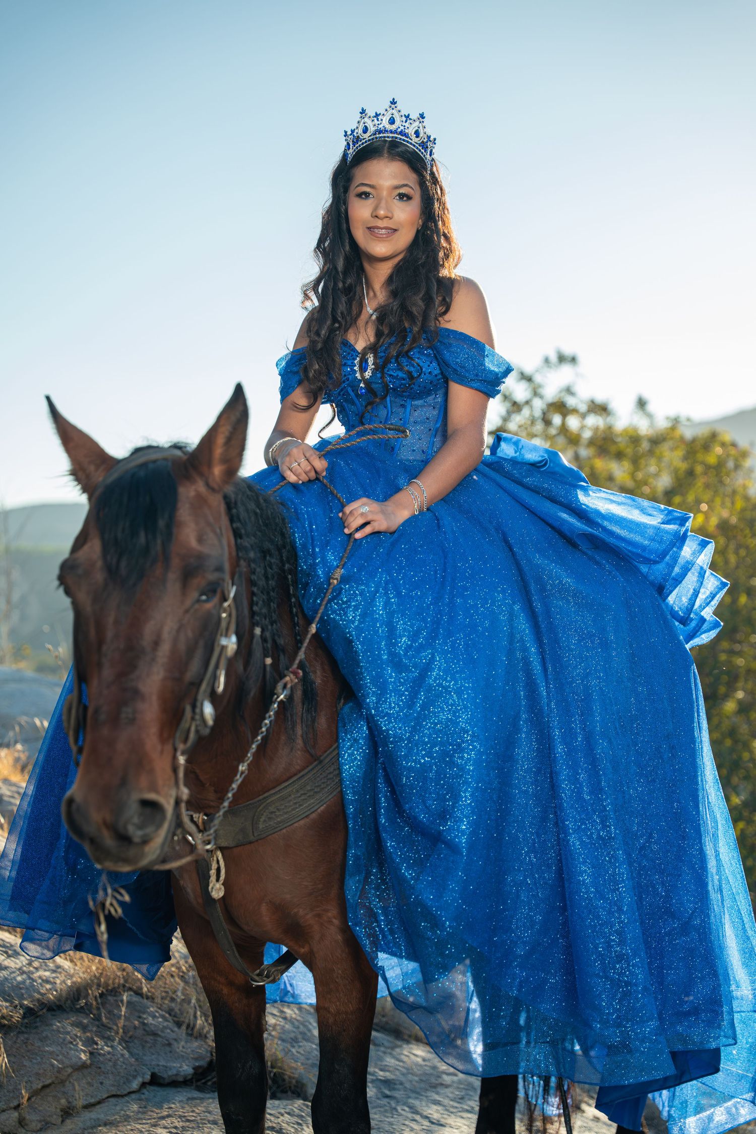 Young quinceañera in a sparkling blue ball gown and crown sits regally on a brown horse outdoors.