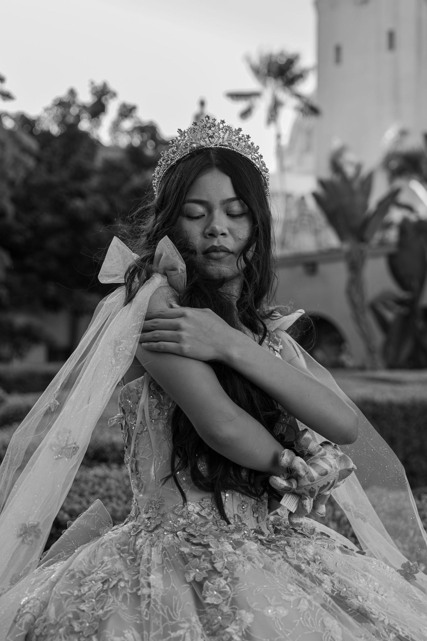 Black and white portrait of quinceañera girl in tiara and floral gown with eyes closed in elegant pose.