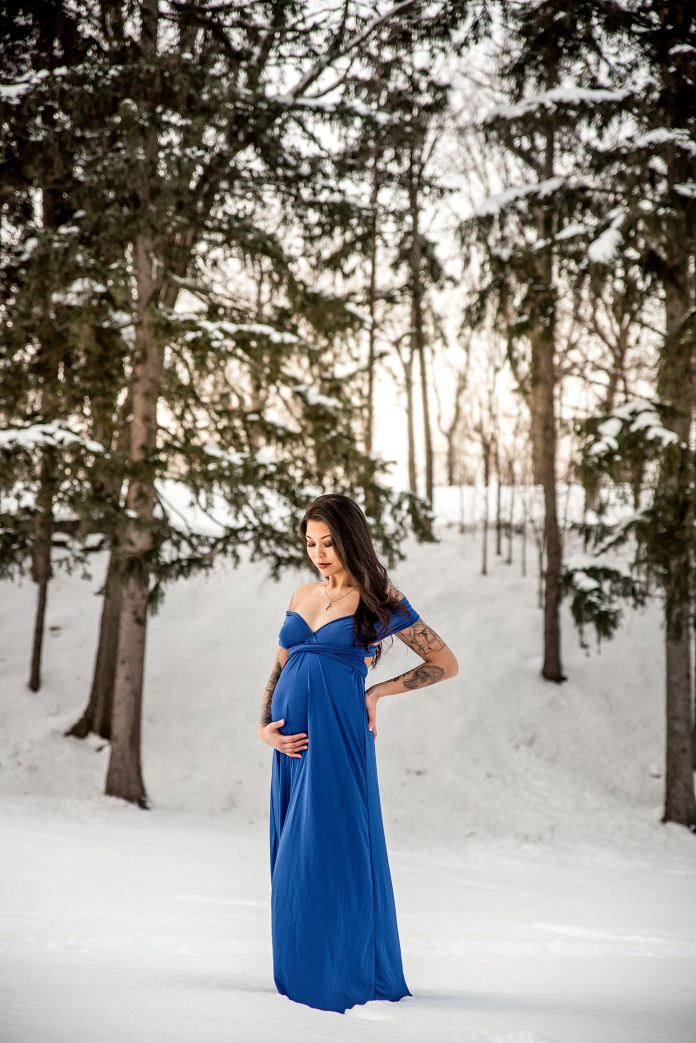 Person in blue dress standing among snow-covered evergreen trees in winter forest.