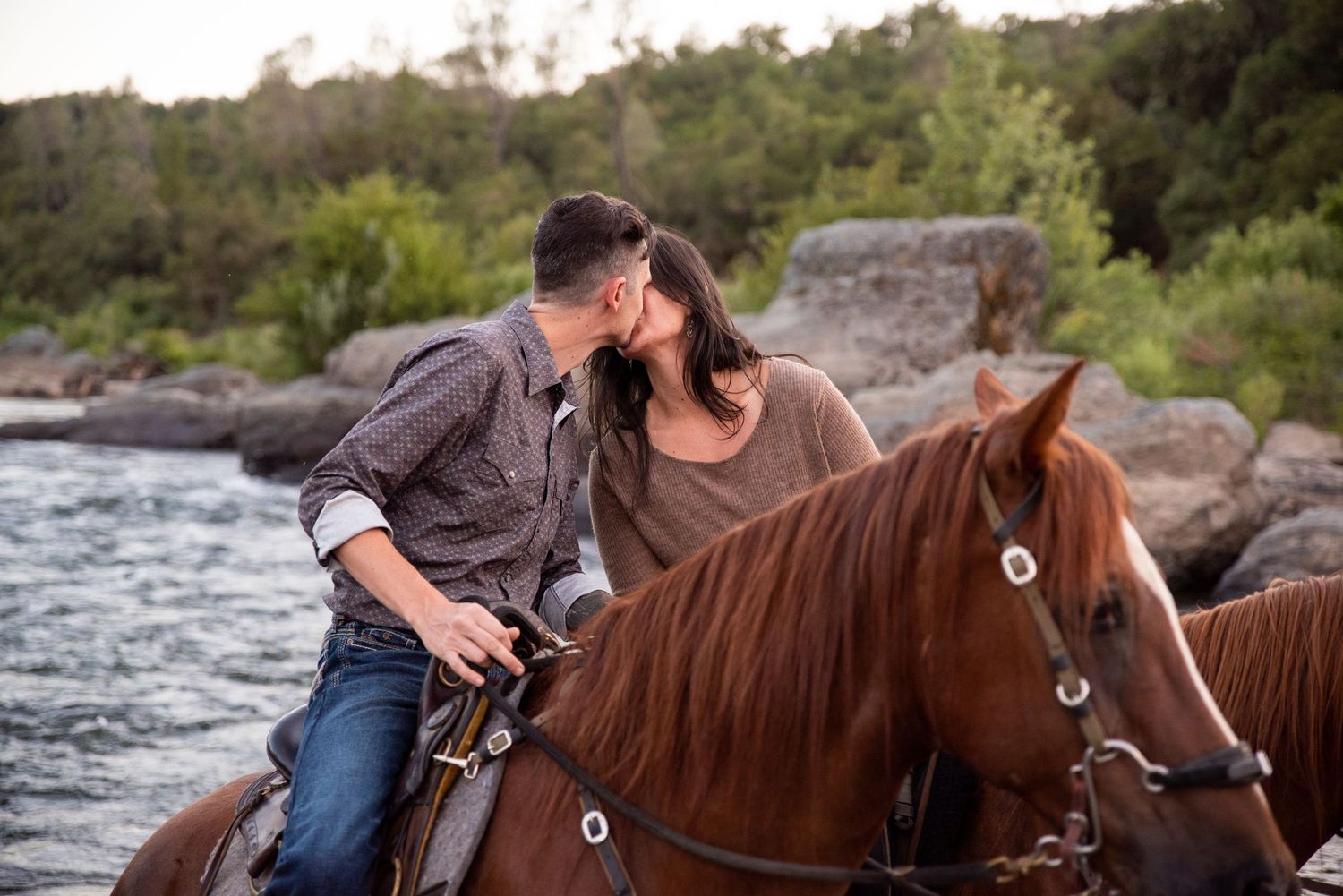 Horseback riders traverse rocky terrain near a river at sunset.