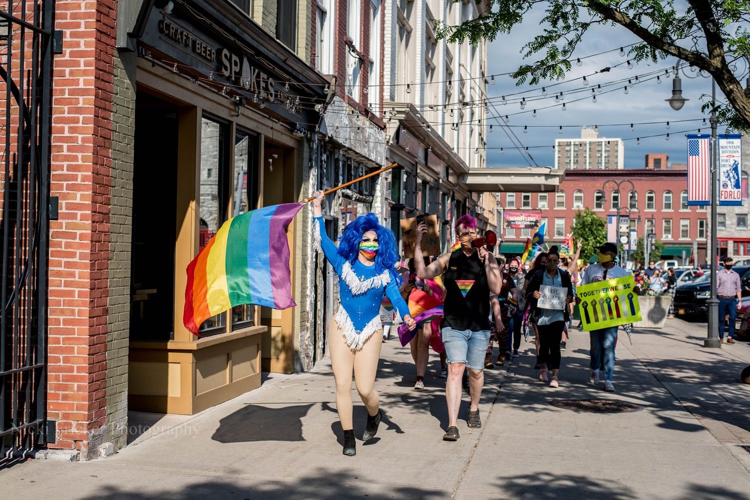 Colorful storefronts and brick buildings line a vibrant downtown street with pride flags and pedestrians.