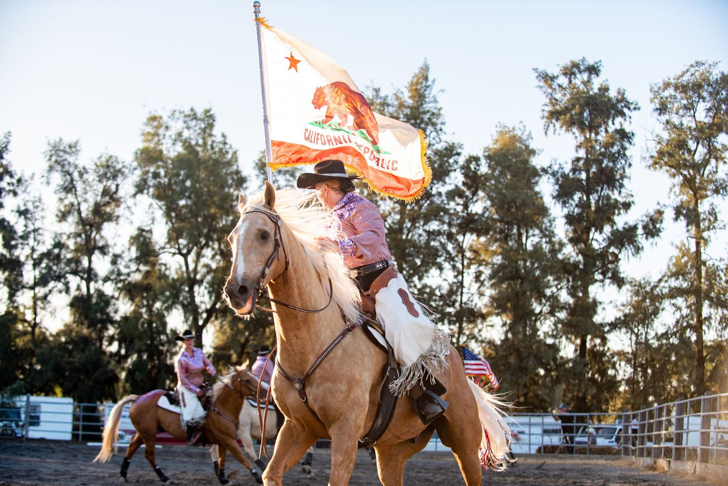 Rodeo riders perform tricks on horseback in an outdoor arena at sunset.
