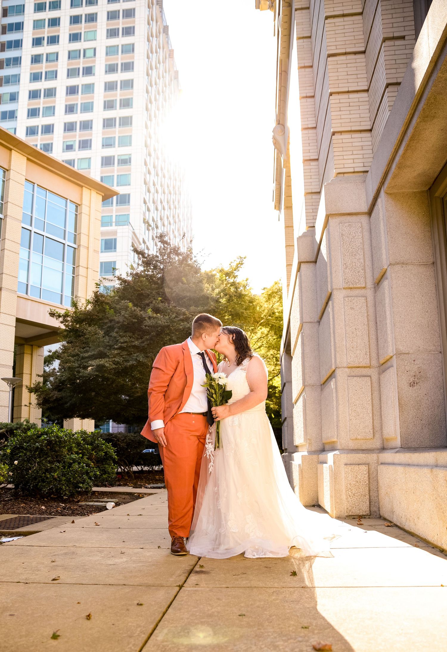 Couple in formal attire embrace near modern office building at sunset.