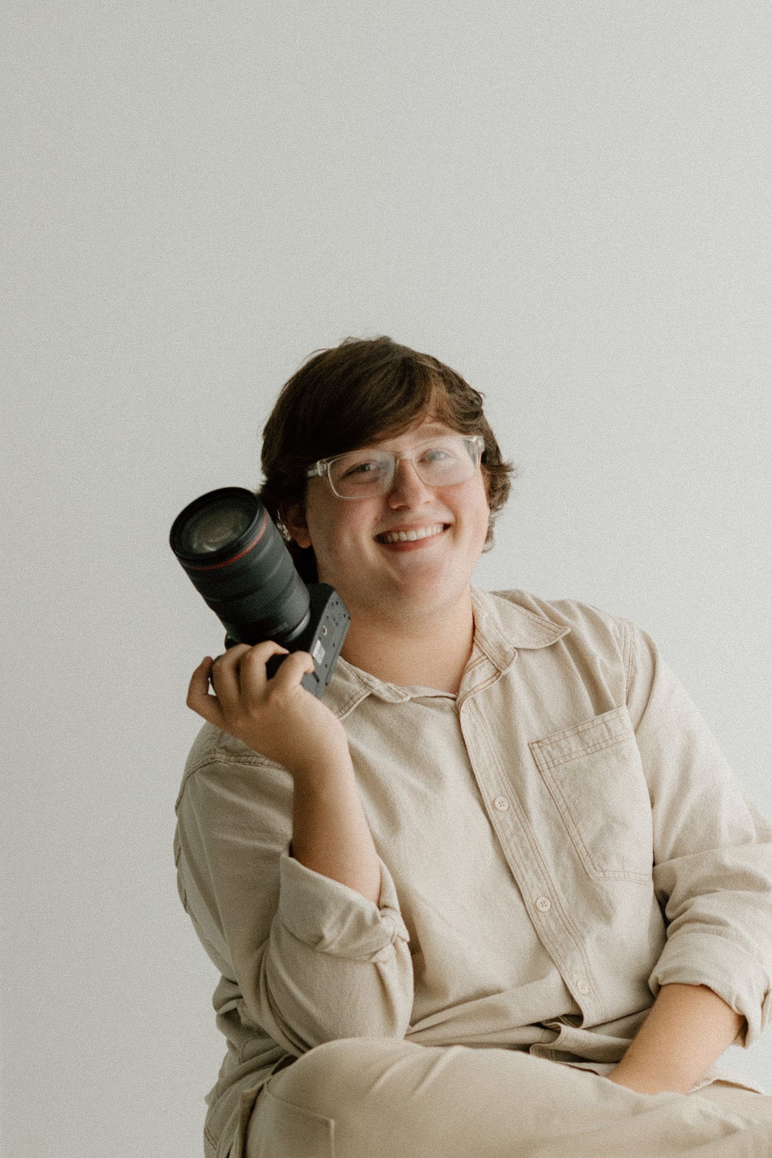 Portrait photographer in beige clothing holding a professional camera against white background.
