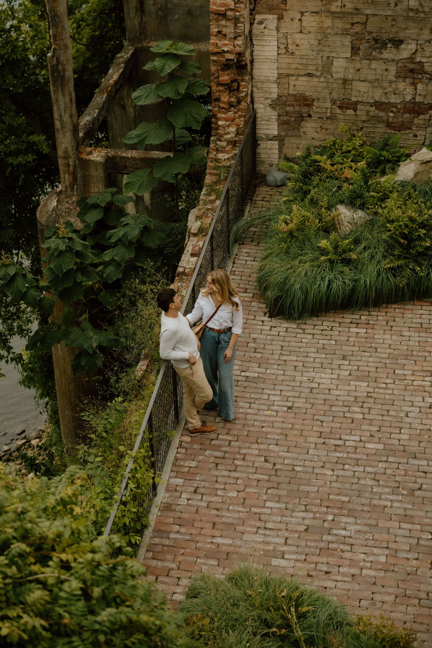 A couple shares an intimate moment against a rustic brick wall surrounded by lush greenery.