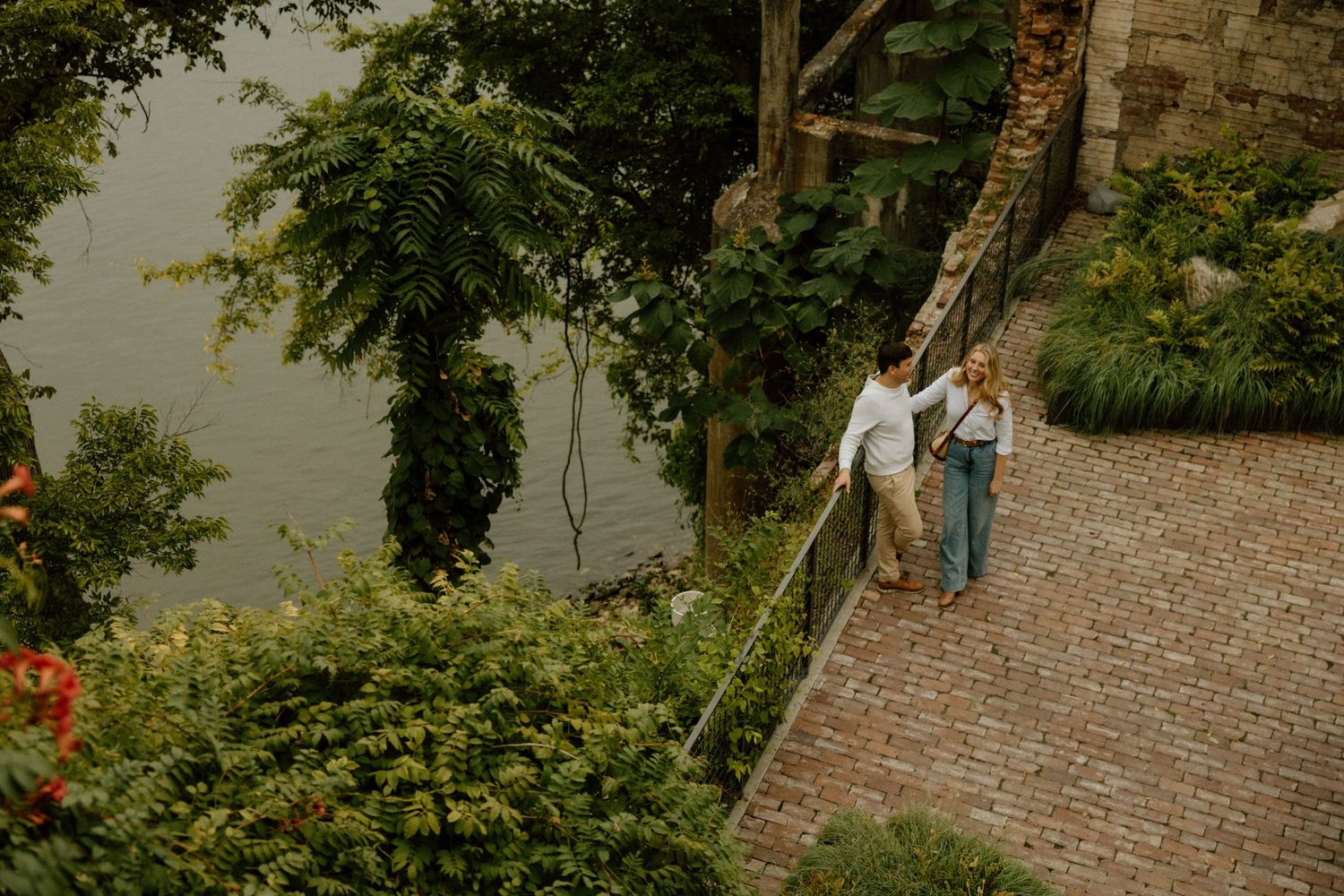 A couple walks down brick steps surrounded by lush landscaping near a waterfront.