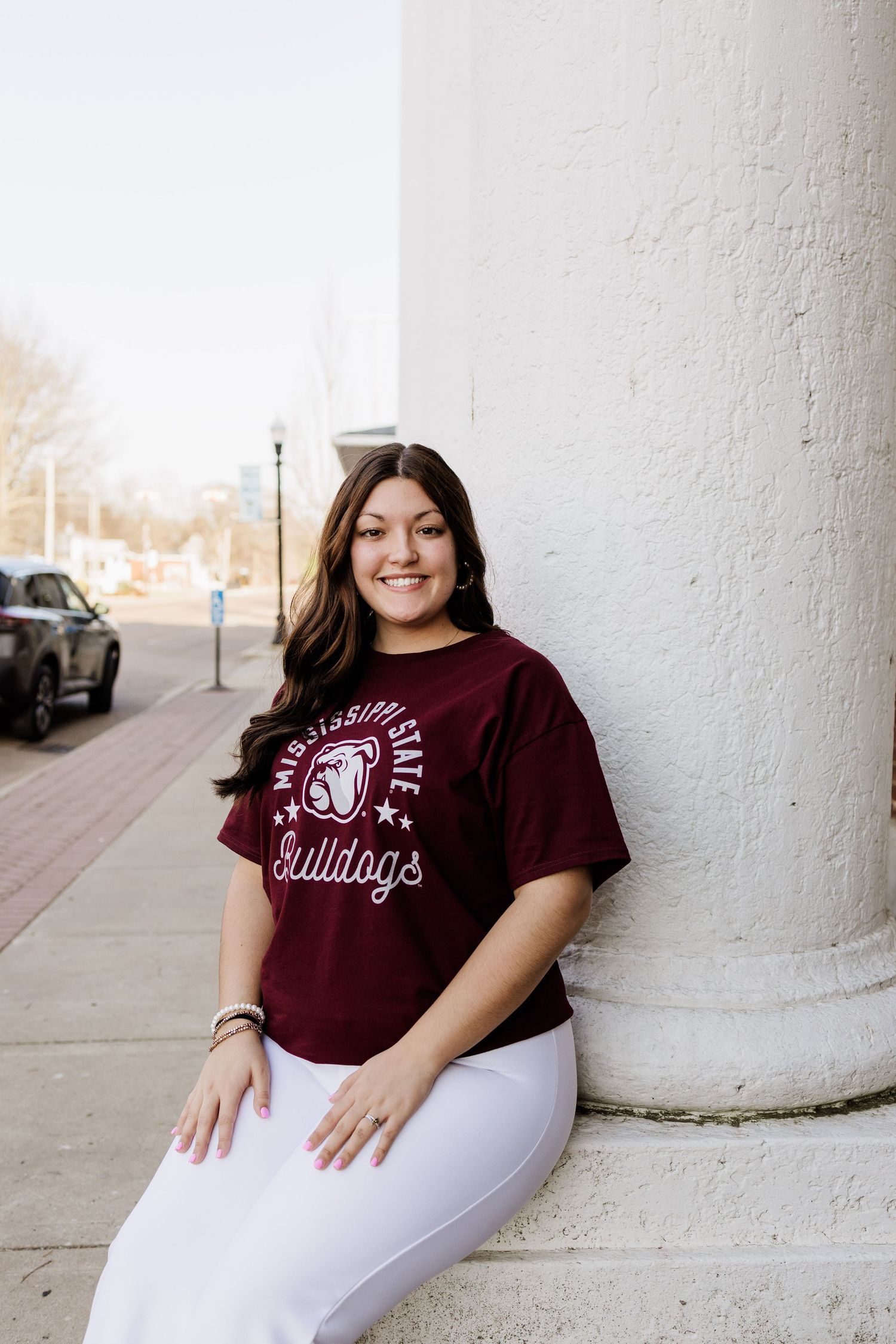 Student in maroon Bulldogs shirt poses against white column on urban street.