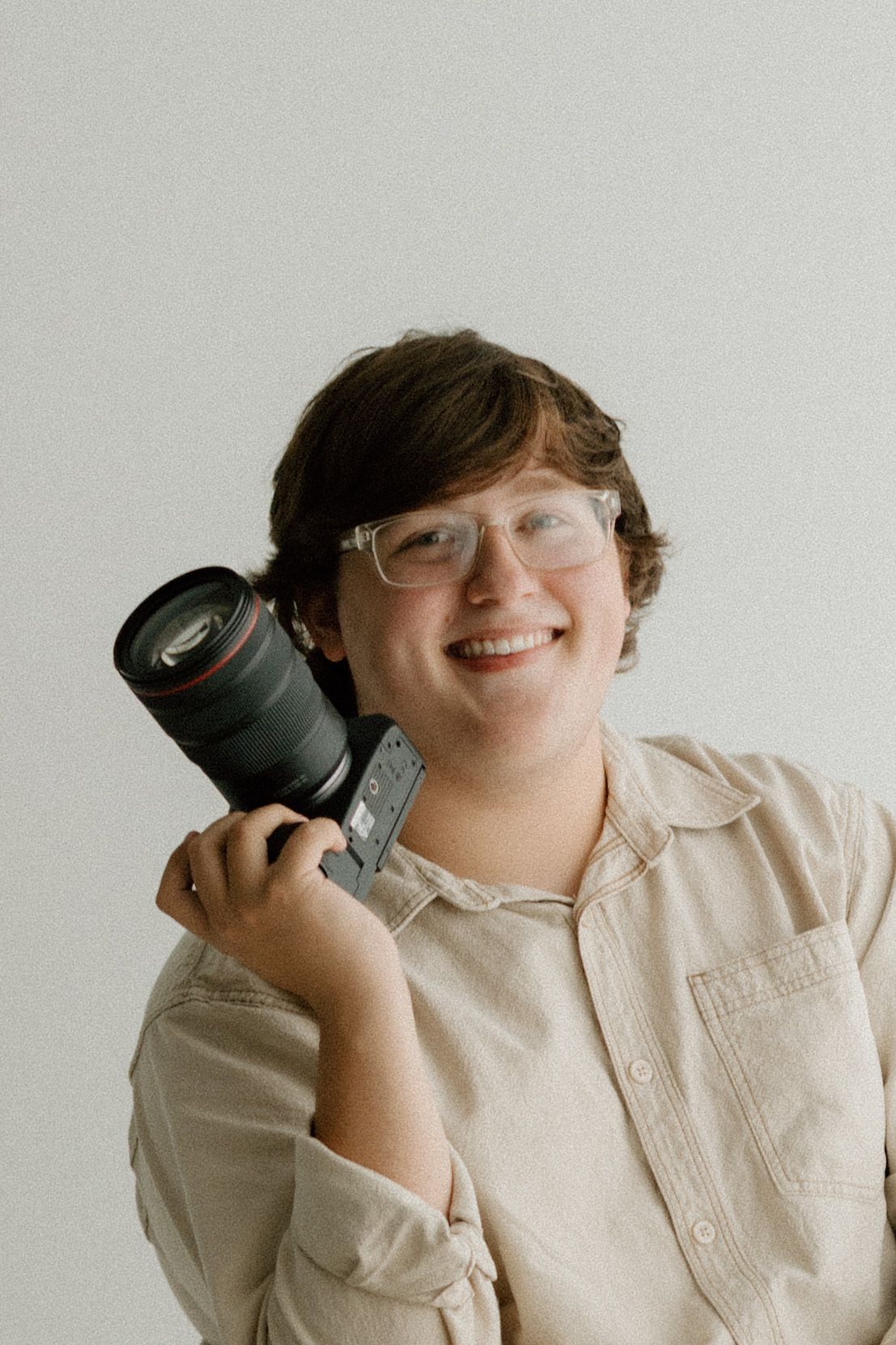 A photographer wearing glasses and a beige button-up shirt holds their camera while smiling.