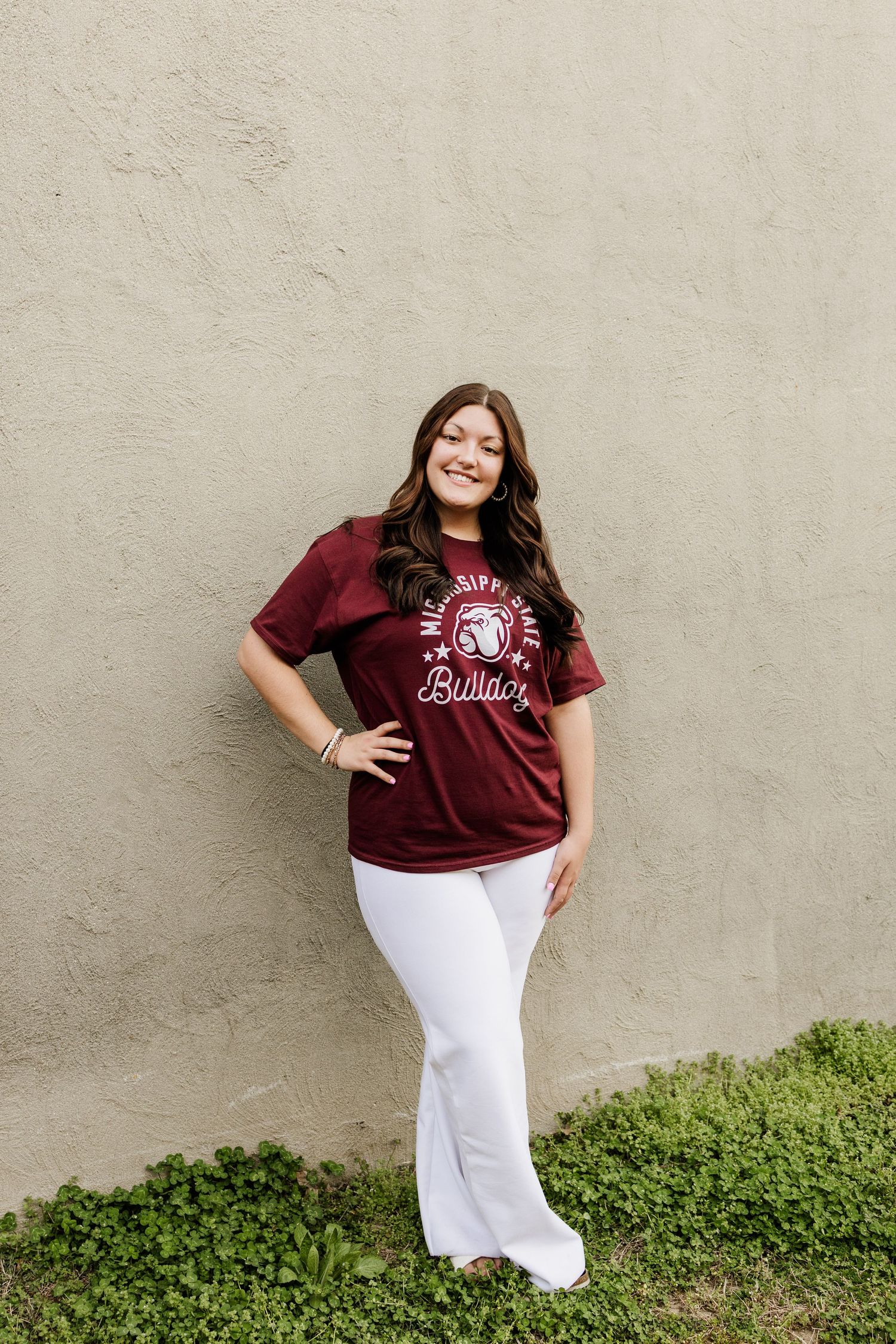 A person in a maroon university shirt and white pants poses against a beige wall.