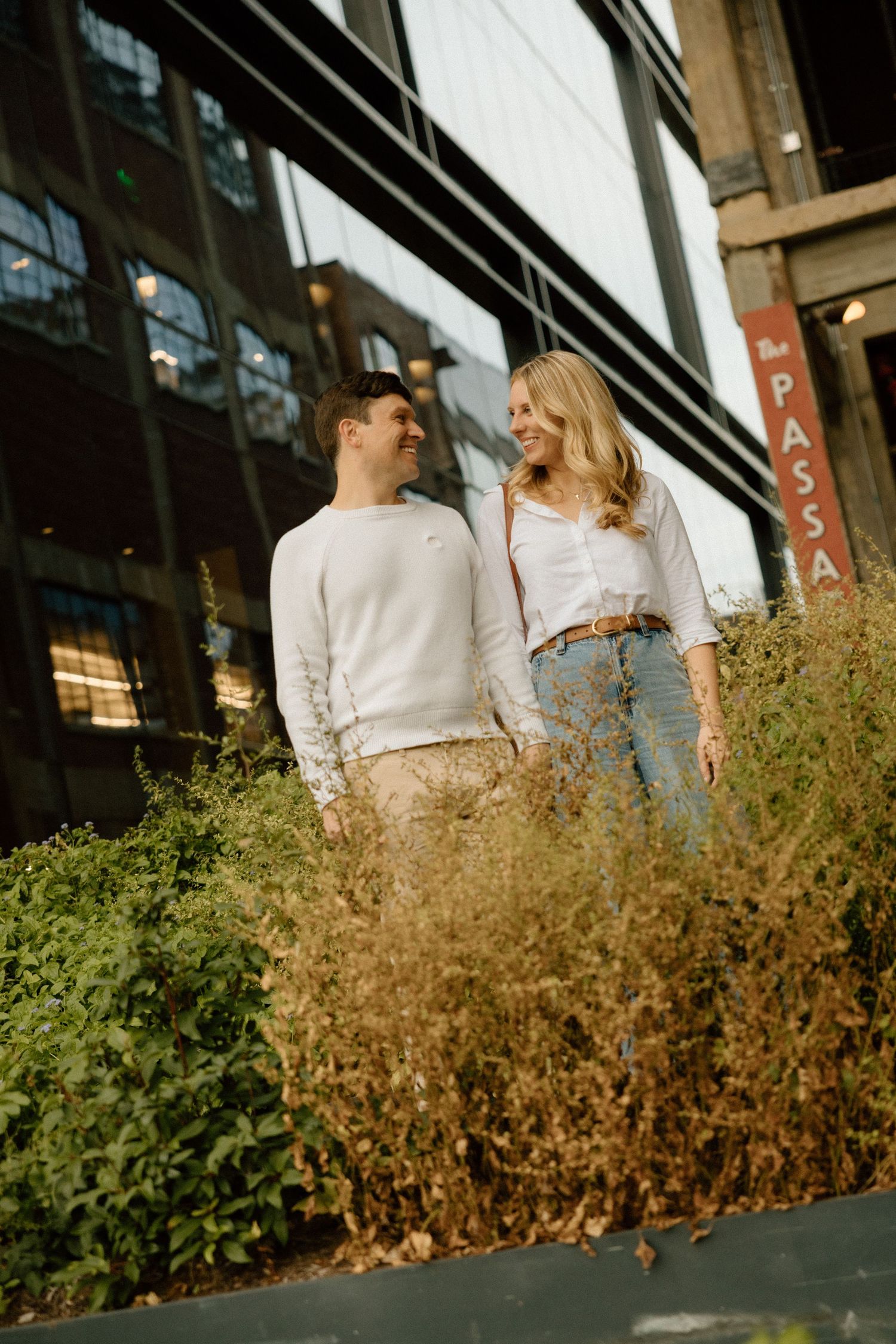A stylish couple stands together near modern architecture with dried grass in the foreground.