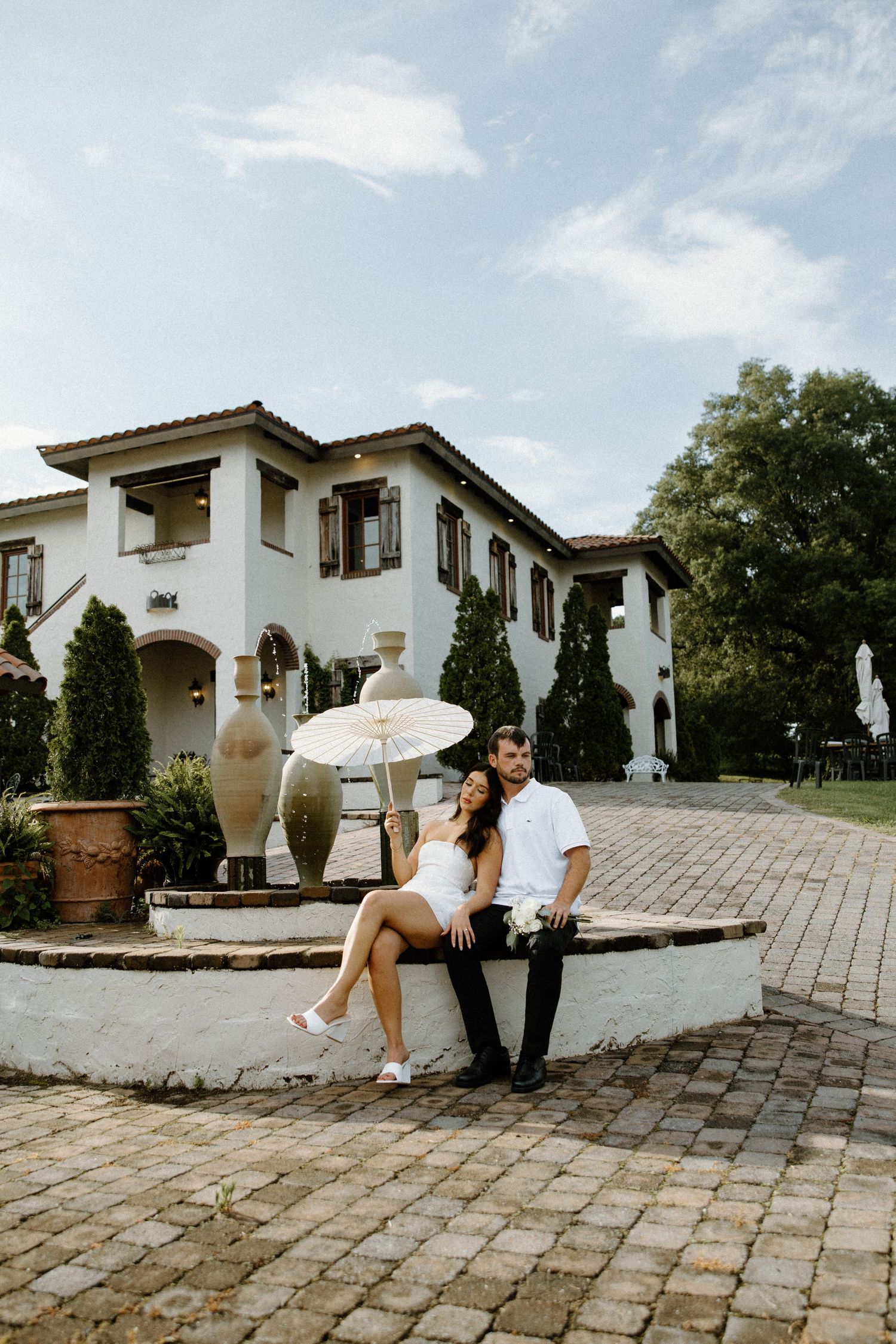 A couple sits together on a white stone fountain outside a Mediterranean-style mansion with brick paving and lush landscaping.