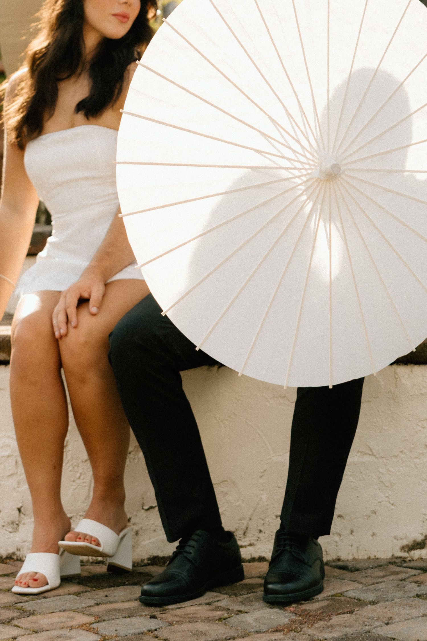 Stylish couple posing with white paper parasol against stone wall.