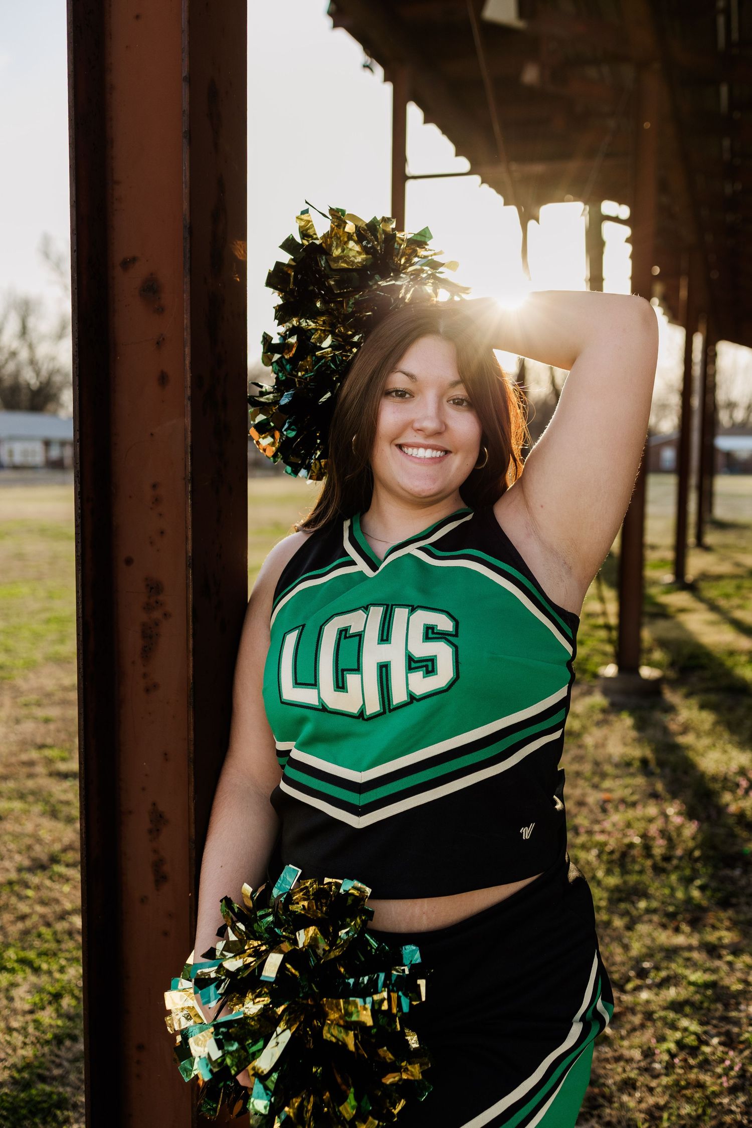 Cheerleader in green uniform poses with pom poms at sunset through pillars.