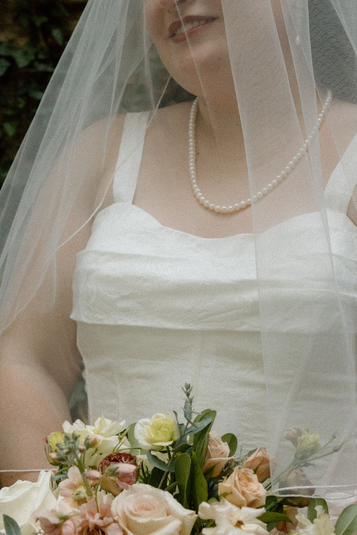 Close-up bridal portrait showing white wedding dress, veil, and pearl necklace with peach and cream bouquet.