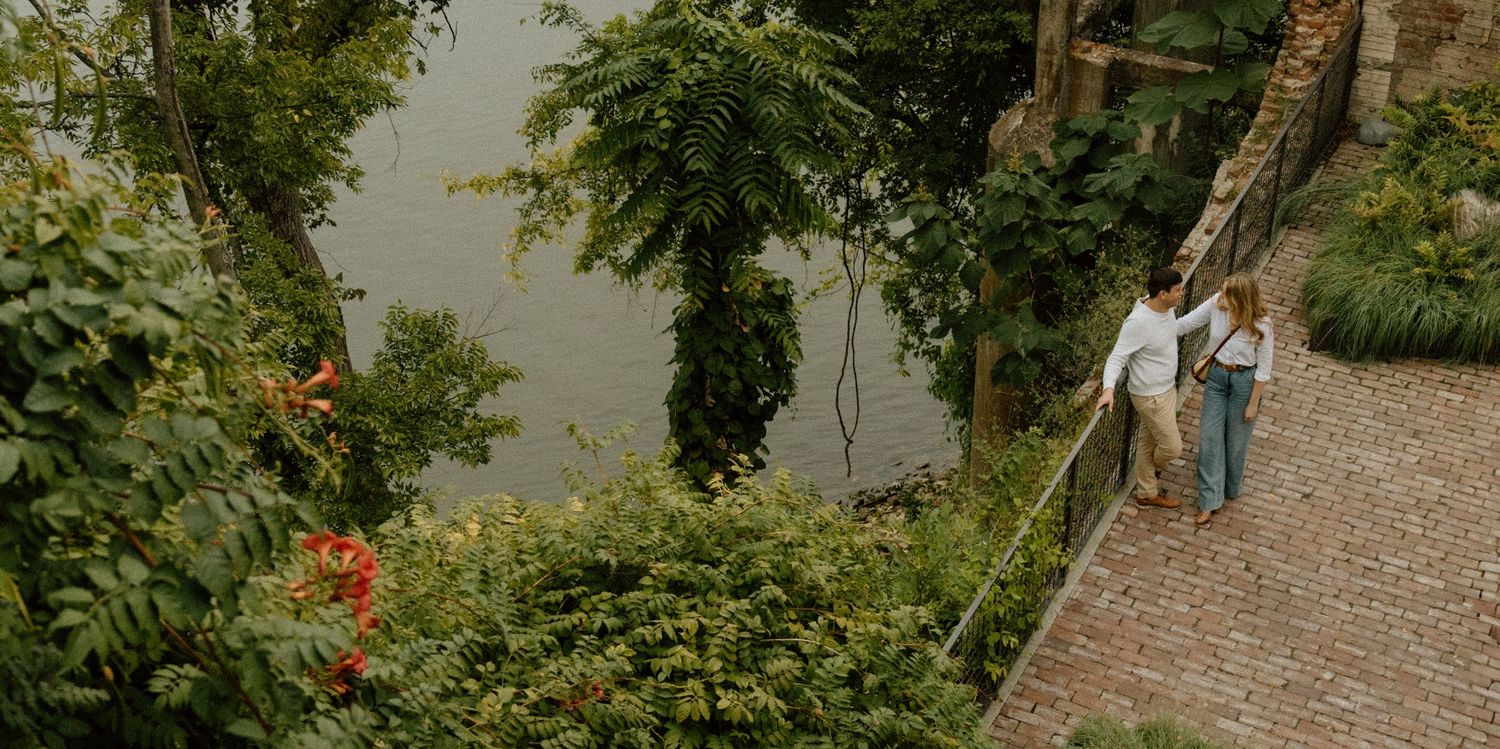 People walking along a tree-lined path by a riverbank on an overcast day.