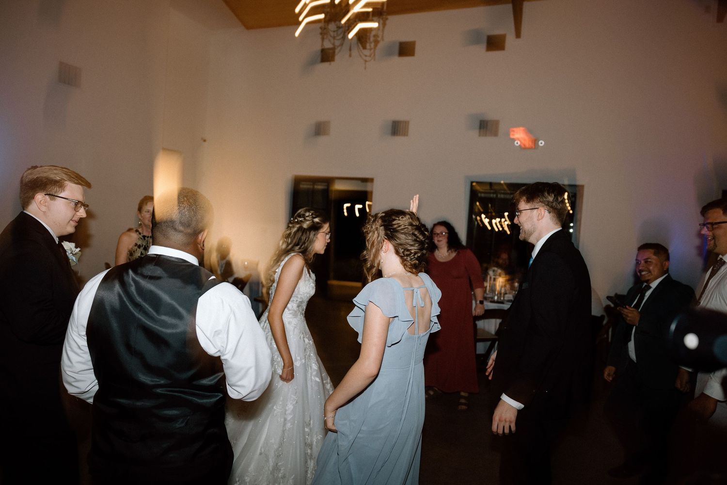 Wedding guests gather on the dance floor during an evening reception in a warmly lit indoor venue.