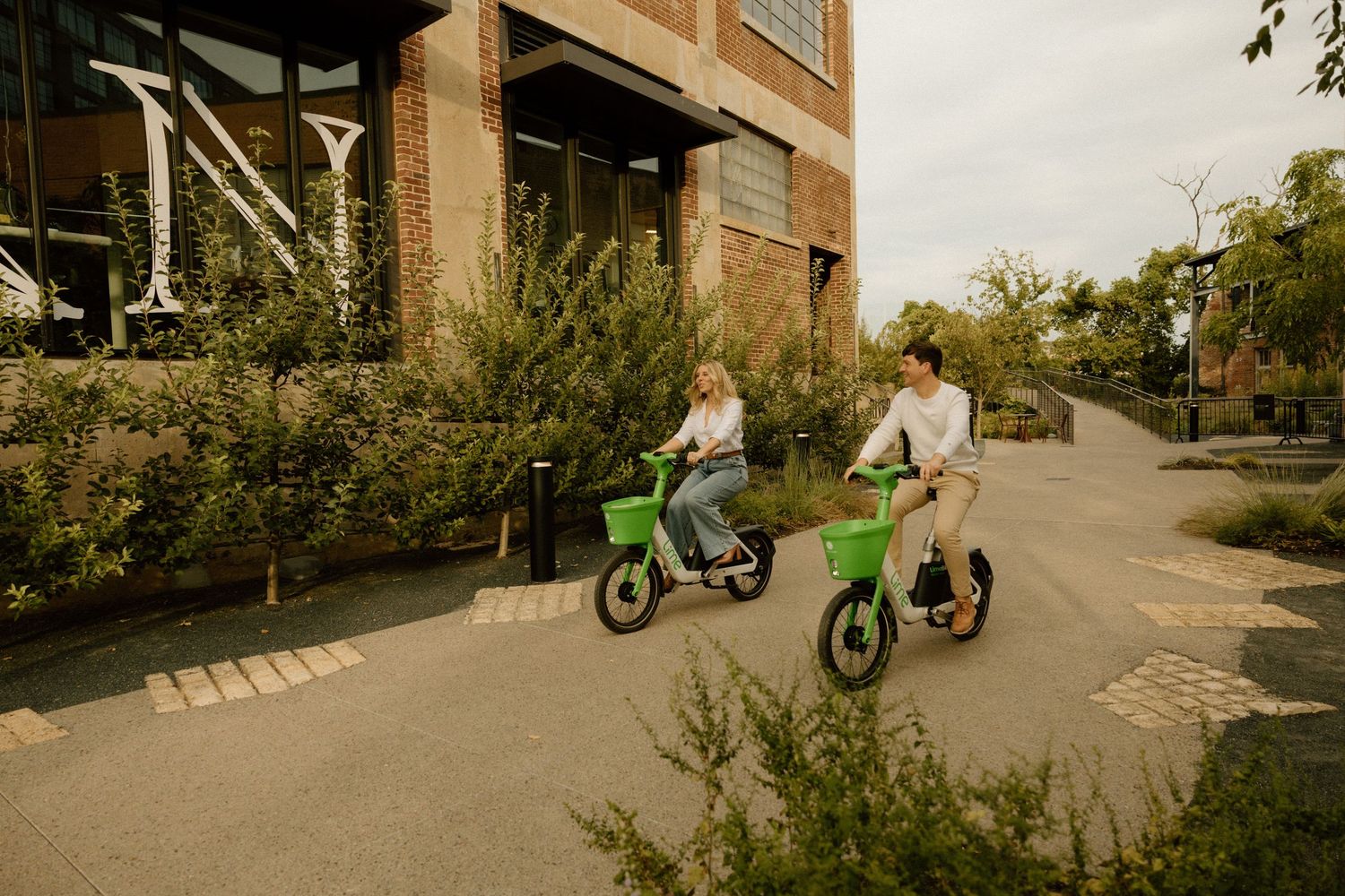 Two people riding bright green scooters along a city street near brick buildings.