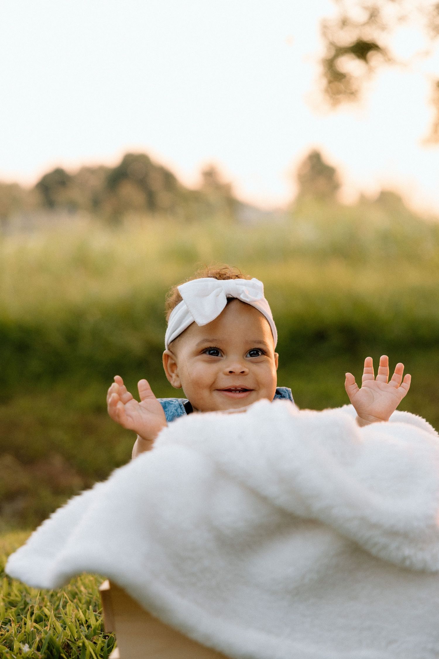 Baby in white bow headband smiling and reaching up.