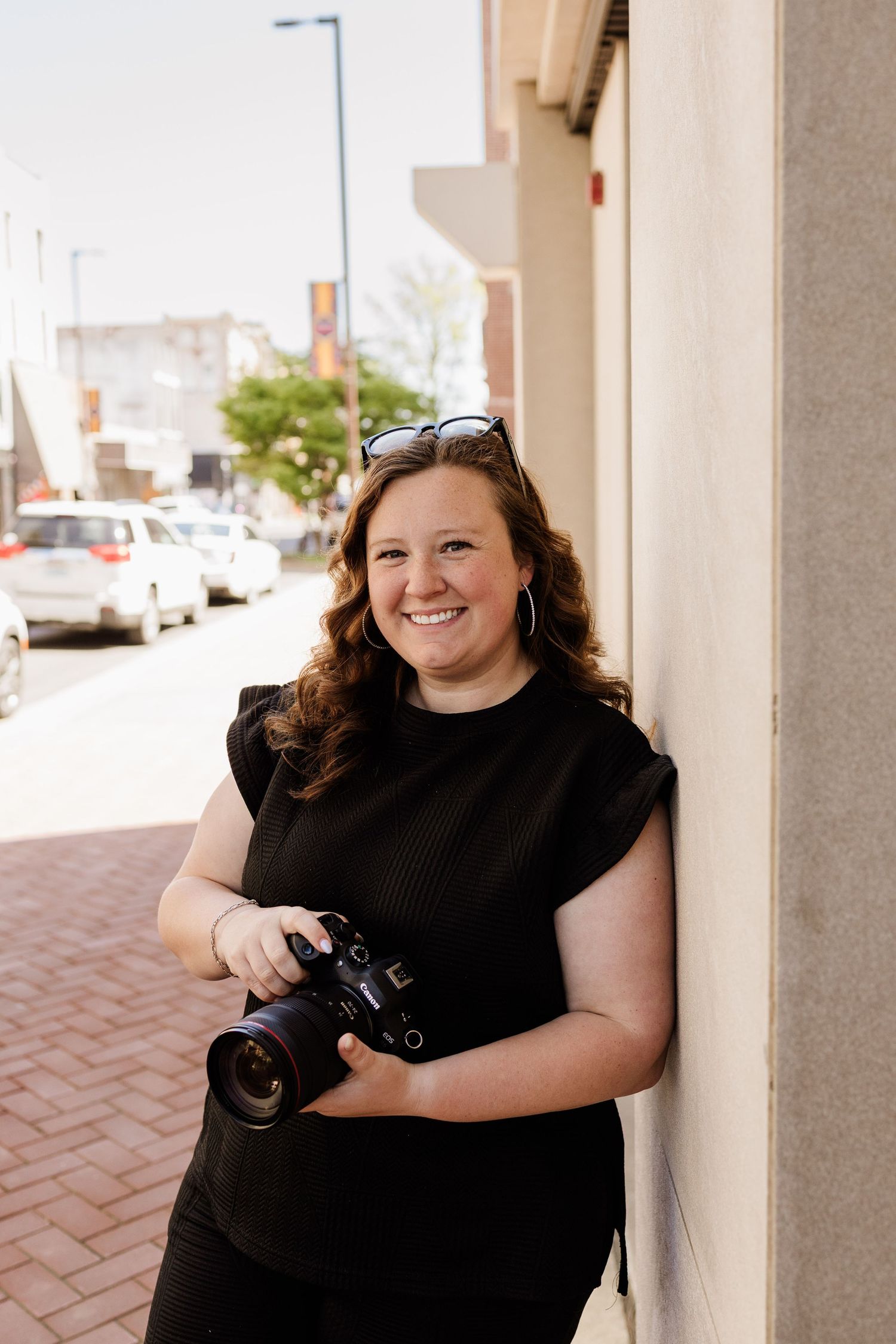 Professional photographer in black attire holds a camera while posing against a wall on a city street.