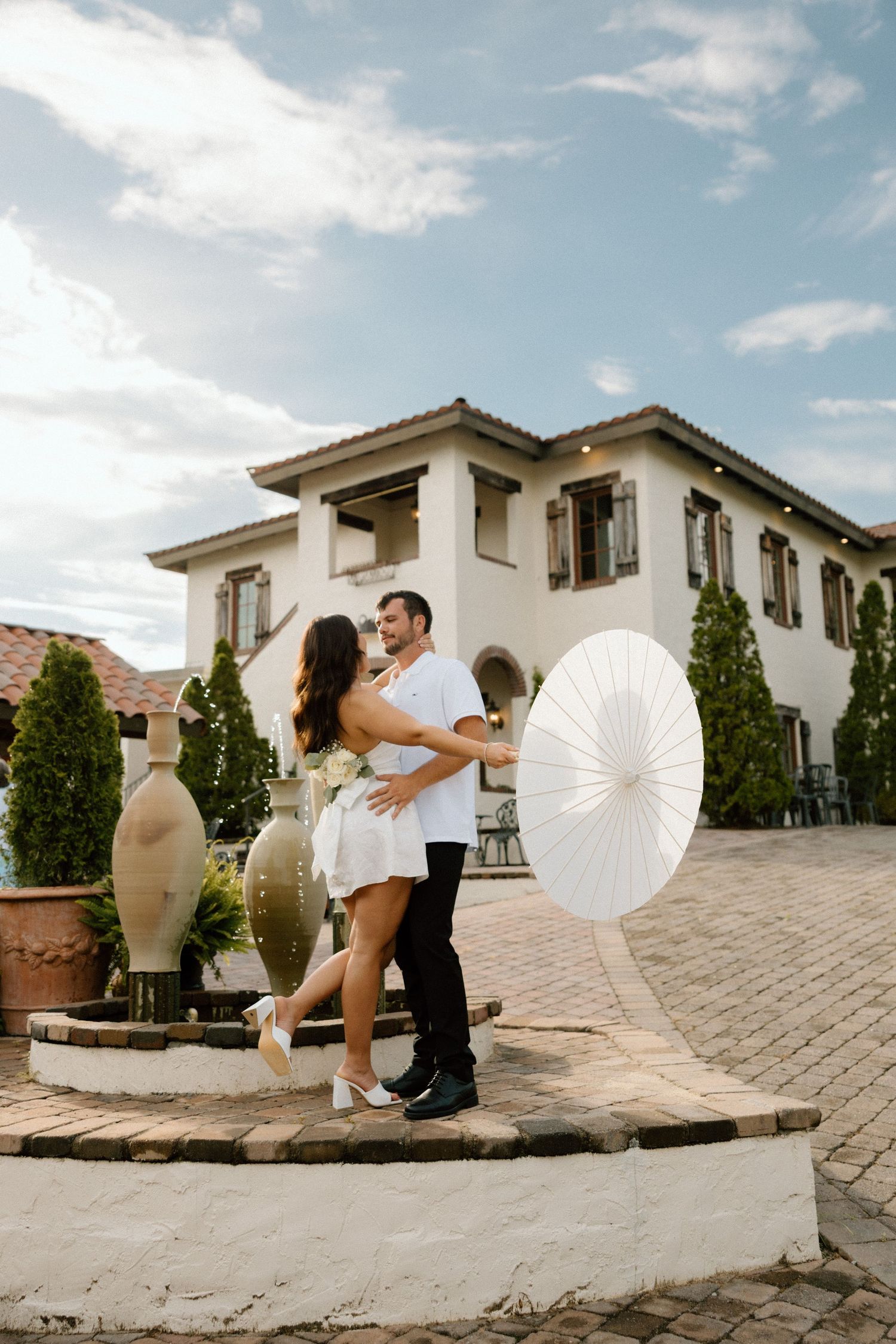 A couple poses with a white parasol against Mediterranean-style architecture.