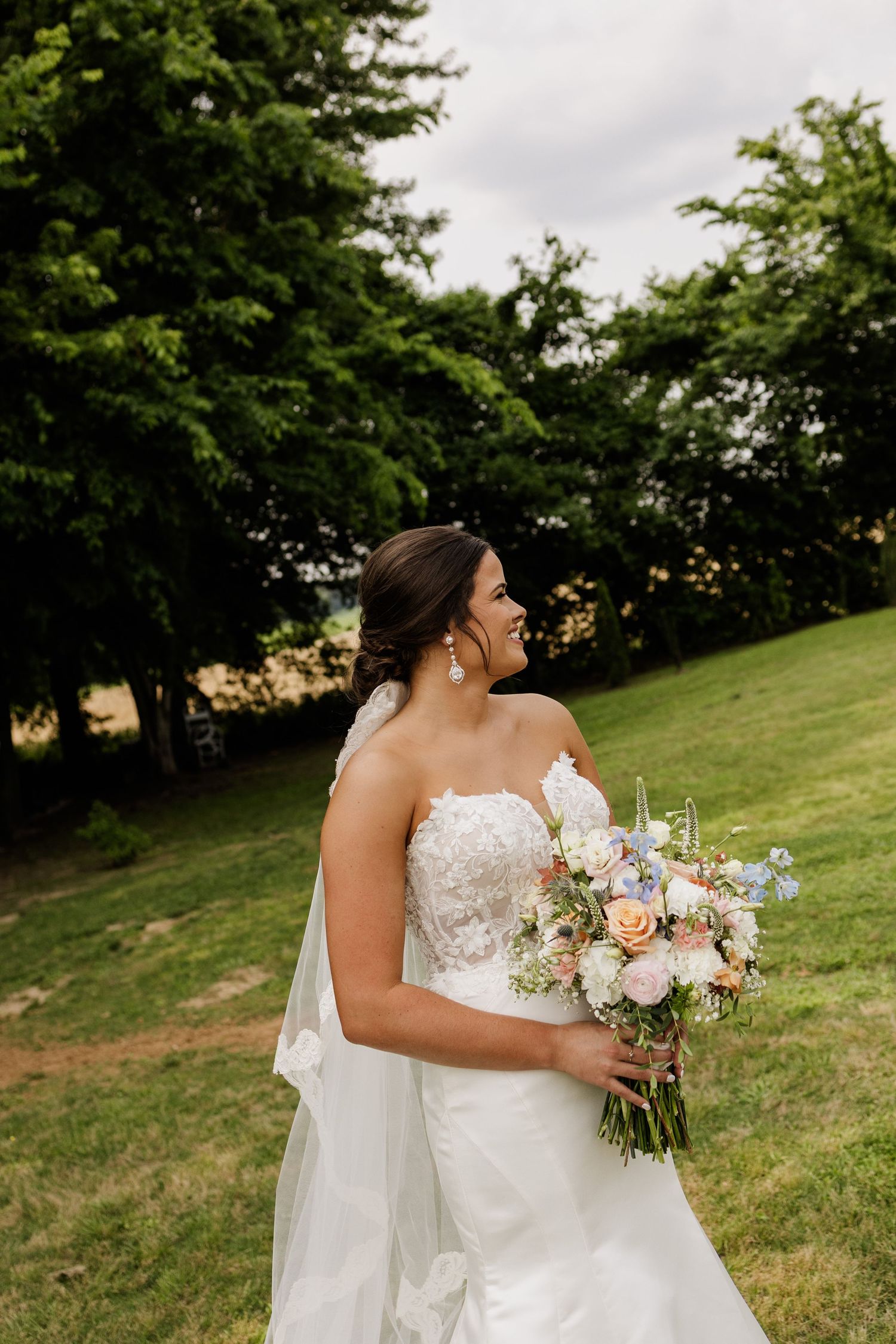 Two people in white wedding dresses embrace while holding floral bouquets on a grassy hillside.