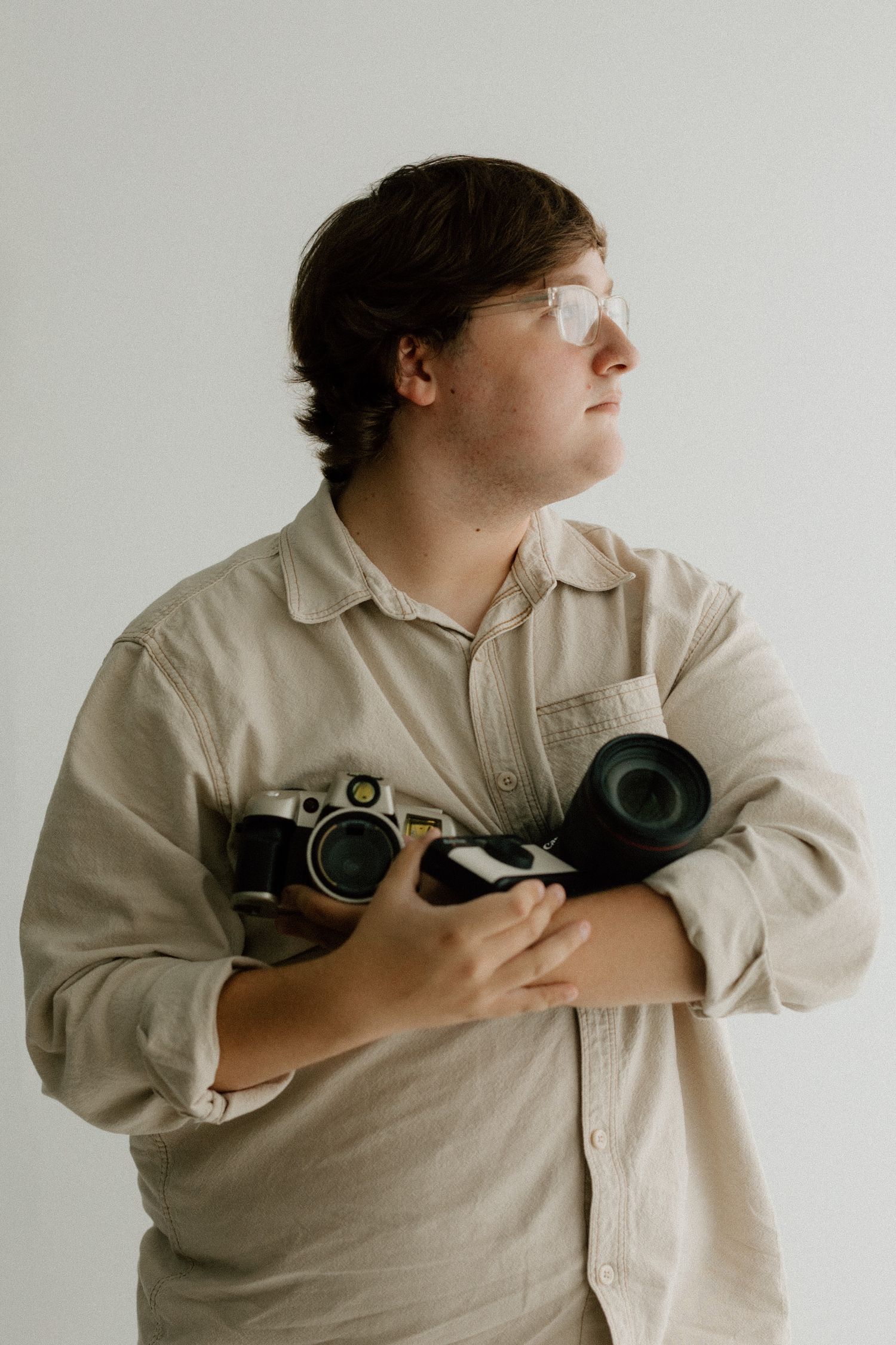 Photographer in beige shirt holding vintage cameras against white background.