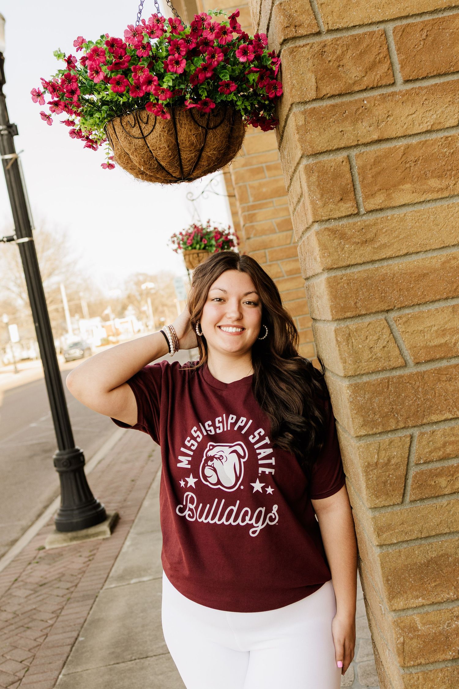 Student in maroon Bulldogs shirt poses beneath hanging flower basket on brick building exterior.