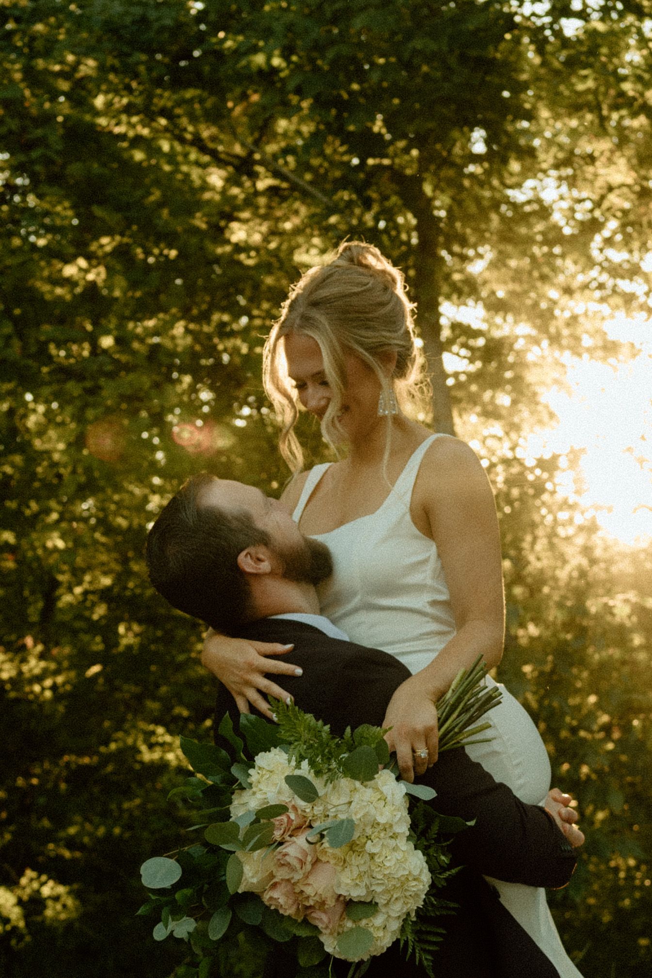 A romantic couple embraces and kisses during sunset in a garden setting with white floral bouquet.