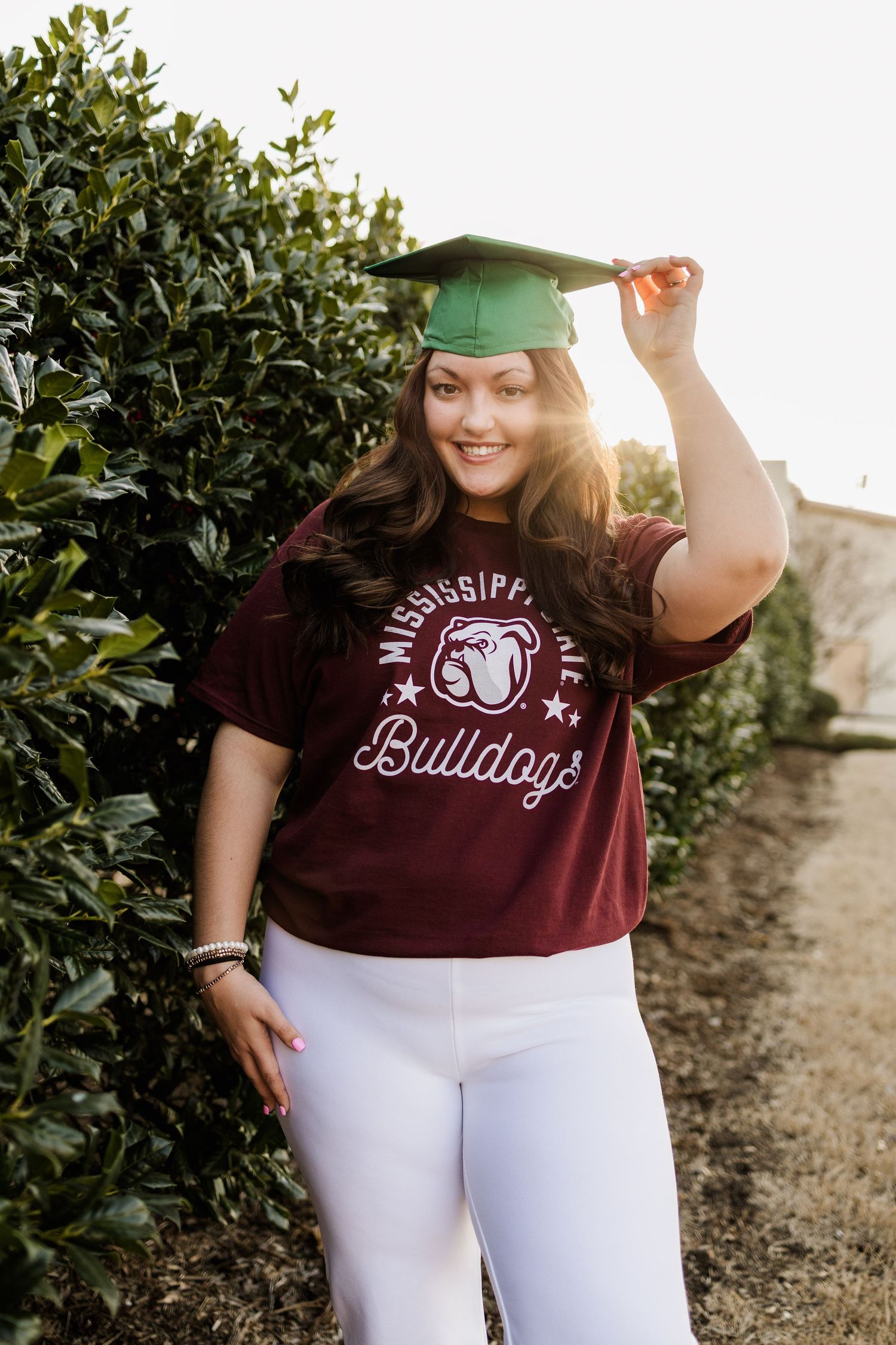 Graduate in maroon Bulldogs shirt and green cap poses by shrubs in natural lighting.