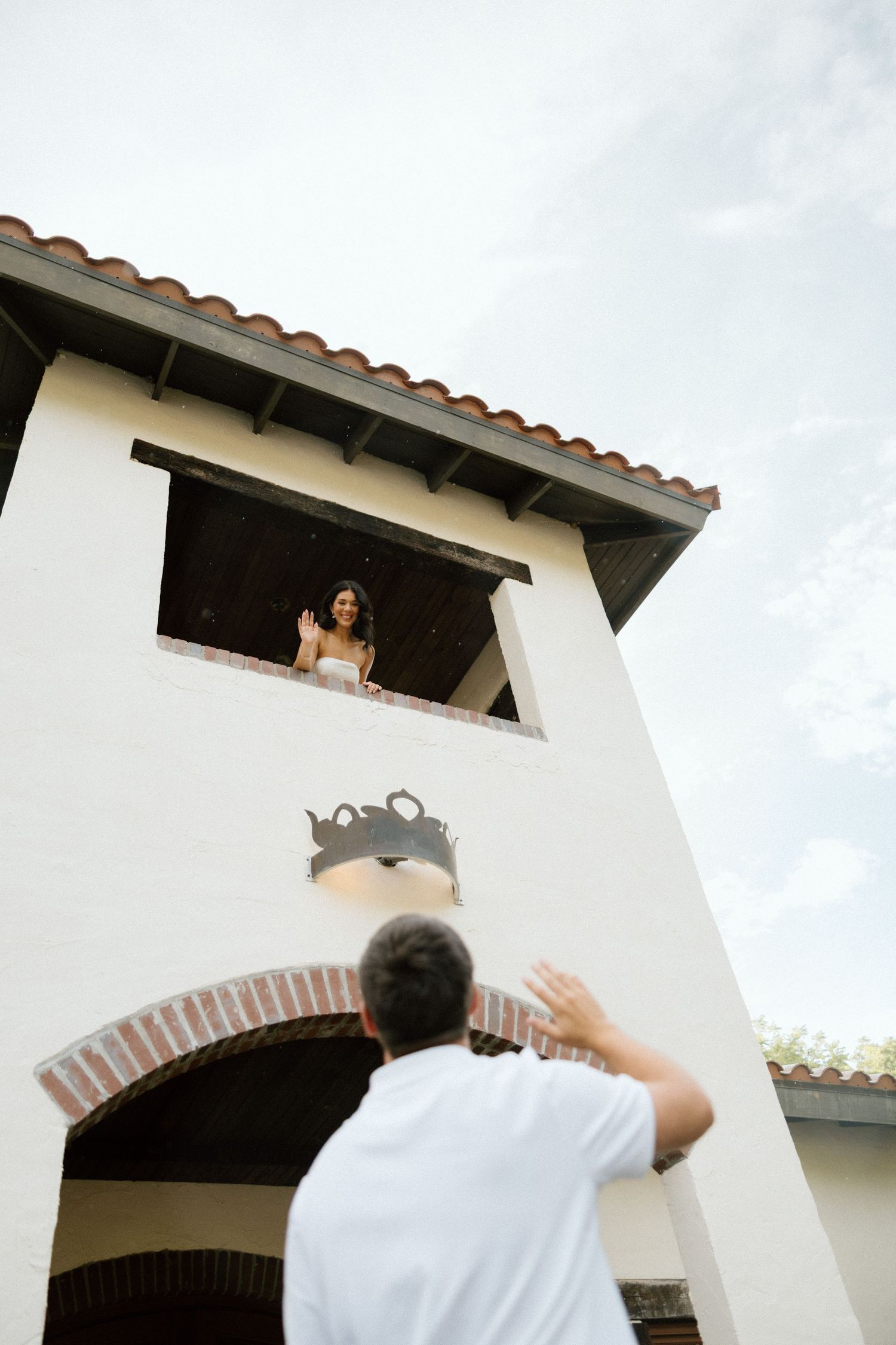 A person in white reaches up toward a Spanish-style building with arched windows and terracotta roof tiles.