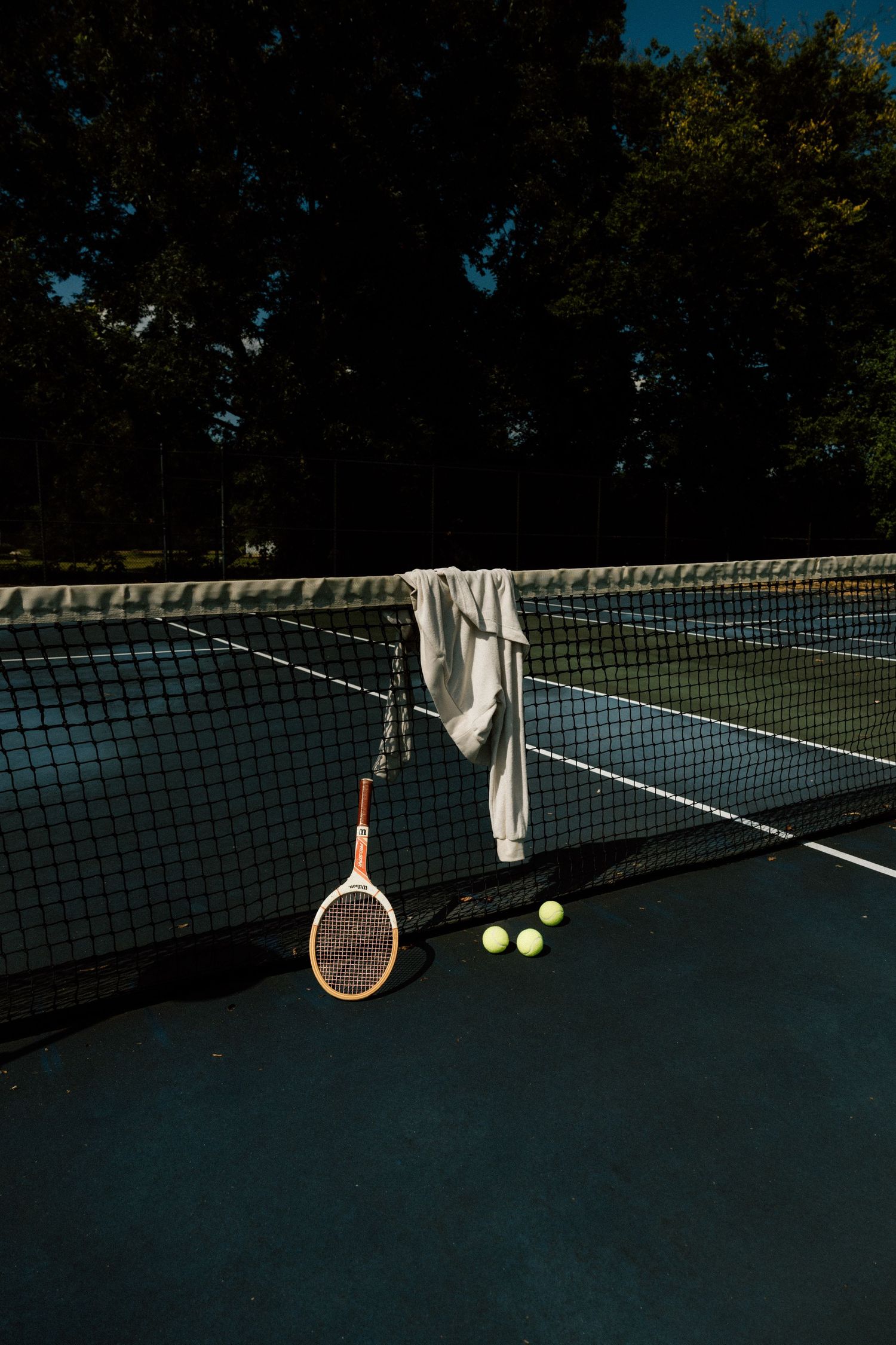 Tennis racket and balls rest against a dark court net at dusk.