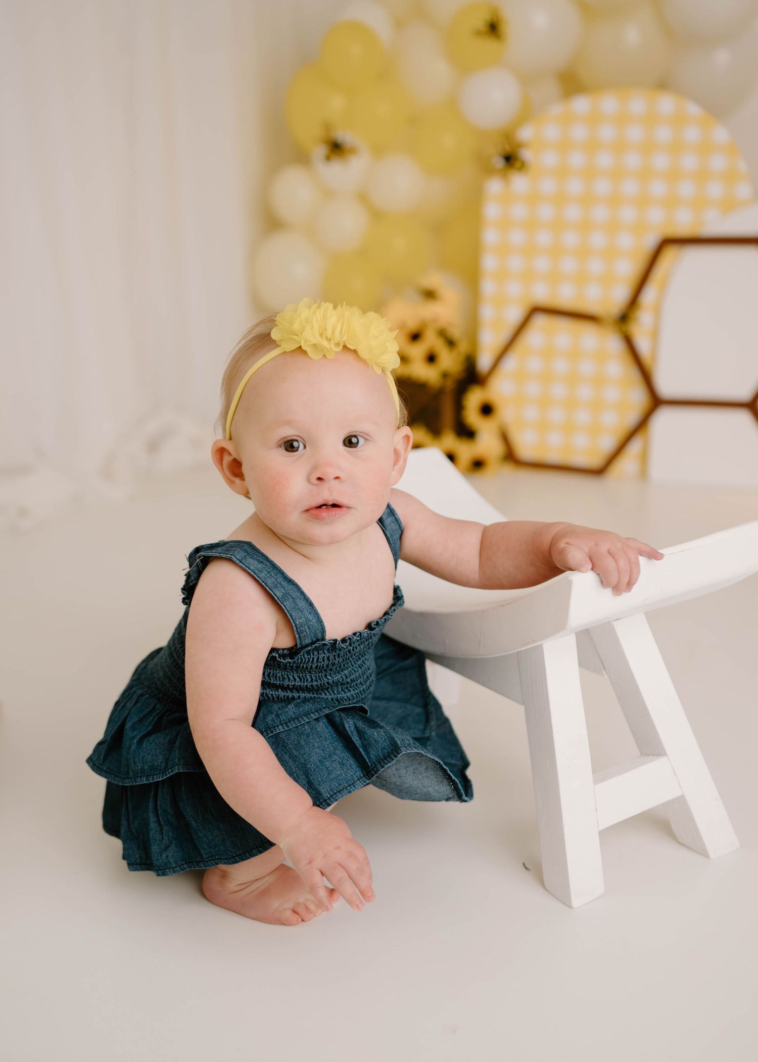 Baby girl in denim dress with yellow bow headband poses by white stool at bee-themed first birthday cake smash.